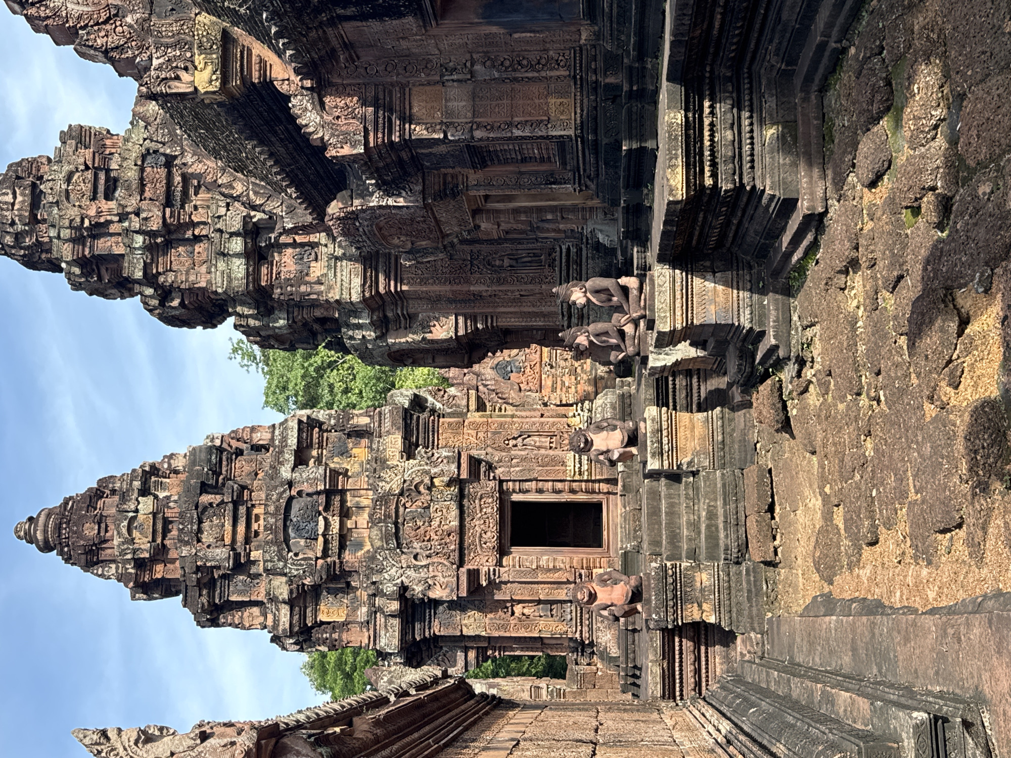 The image shows the central sanctuary of Banteay Srei temple in Cambodia, a Hindu temple dedicated to Shiva. The temple is constructed of red sandstone and features intricate carvings and bas-reliefs. The central sanctuary consists of a series of towers, or prasats, which are tiered and decorated with mythical figures and floral motifs. In the foreground, four stone statues of mythical creatures, possibly lions or guardian deities, are placed on pedestals. The ground in front of the sanctuary is paved with rough stones. The sky is blue with scattered clouds, and lush green trees are visible in the background. The overall impression is one of ancient grandeur and religious significance.