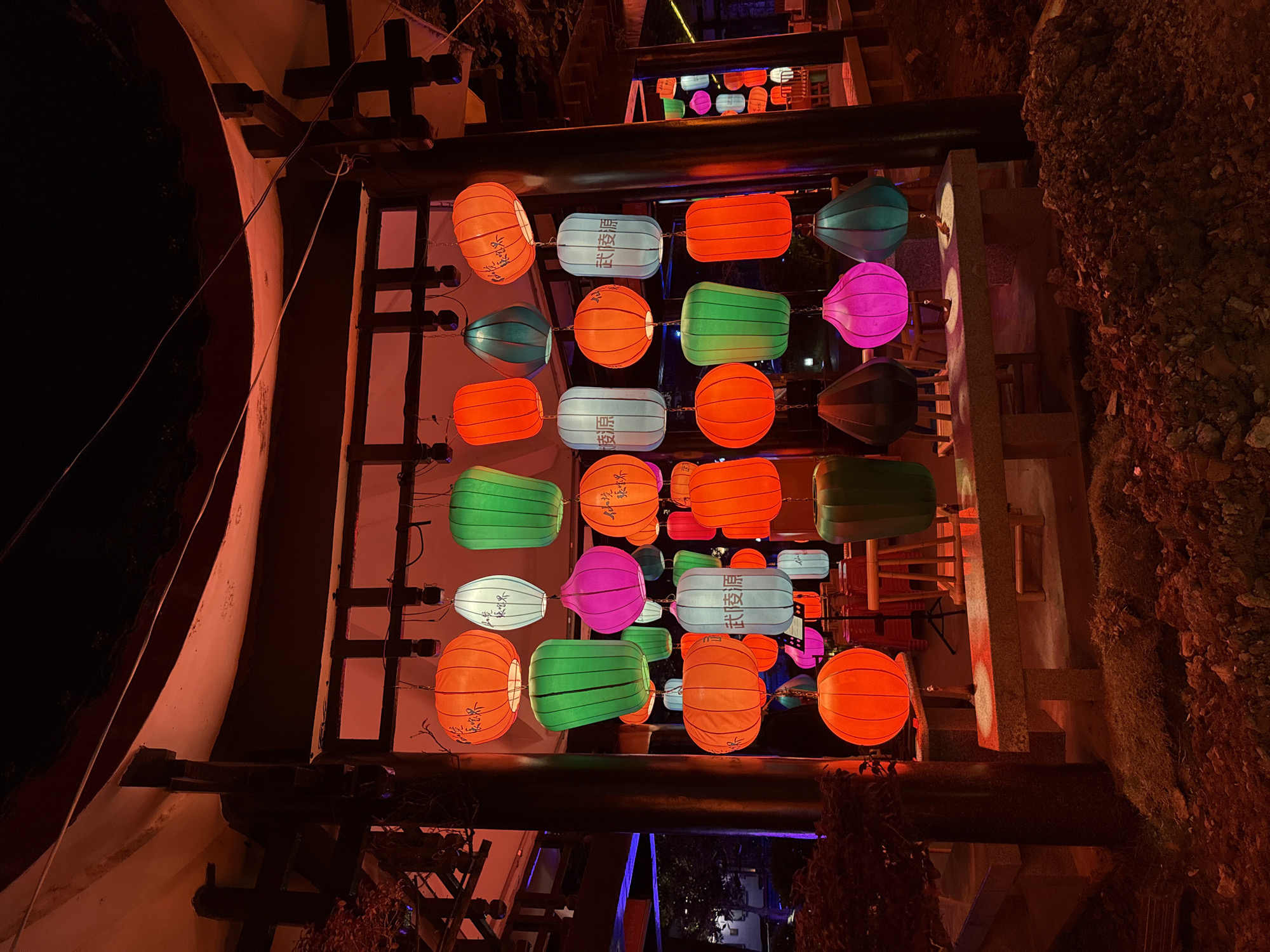 The image displays a vibrant night scene in Wulingyuan, China, showcasing a decorative array of traditional Chinese lanterns. The lanterns are arranged in a grid pattern, suspended from dark wooden beams, and brightly illuminated from within. They come in various shapes, including spherical, oval, cylindrical, and rectangular, and are colored in striking shades of orange, light blue, green, and magenta/pink.
A notable detail is the visible Chinese text on some of the light blue cylindrical lanterns, clearly spelling "武陵源" (Wǔlíngyuán), which confirms the specified location. Other lanterns, particularly the orange and green ones, feature delicate calligraphic script, though the specific characters are not clearly legible.
The scene suggests an evening or night setting due to the overall darkness and the prominent illumination from the lanterns. Architectural elements such as dark wooden beams and structural supports are visible, framing the lantern display. To the left, a light-colored curved wall or structure partially frames the view, while to the right, a rugged, rocky embankment rises, suggesting a natural or mountainous backdrop. The display creates a festive and culturally rich atmosphere, likely part of an outdoor event or permanent decoration.