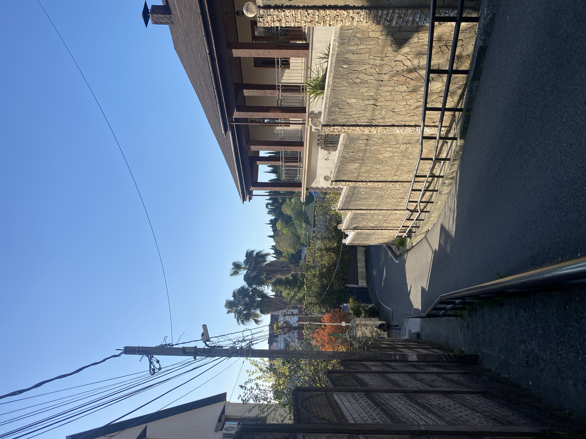 The image, which appears to be rotated 90 degrees counter-clockwise, depicts a sunny residential street scene in Sochi, Russia, characterized by lush greenery and varied architecture.
In the foreground and mid-ground, on the right side of the frame, a multi-story building features a dark brown tiled roof and walls clad in textured, light-brown stone. Several balconies with ornate dark metal railings extend from the building, supported by robust dark pillars. Green plants are visible in planters on these balconies, and a chimney with a decorative cap protrudes from the roofline against a clear blue sky.
An asphalt road curves downwards and to the left through the center of the image. It is bordered on the right by a stone retaining wall and a black metal guardrail that follows the curve of the street. On the left side of the foreground, a dark, textured fence or gate runs parallel to the viewer, and behind it, part of a lighter-colored building with an air conditioning unit is visible. A prominent utility pole with a dense tangle of overhead wires stands in the mid-ground on the left, with additional wires stretching across the clear blue sky.
The background and mid-ground are rich with vegetation, including several large palm trees and other deciduous and evergreen trees, contributing to the verdant appearance. One tree in the mid-ground on the left displays striking reddish-orange foliage. In the distance, other residential structures with light-colored walls and reddish roofs are nestled amidst the dense greenery. A lone person can be seen walking on a path or sidewalk in the mid-ground on the left, near the utility pole.
The scene is bathed in bright sunlight under a clear, cloudless blue sky, casting distinct shadows that suggest either morning or late afternoon. The overall impression is one of a serene and verdant residential area in a warm climate. No visible text is present in the image.
