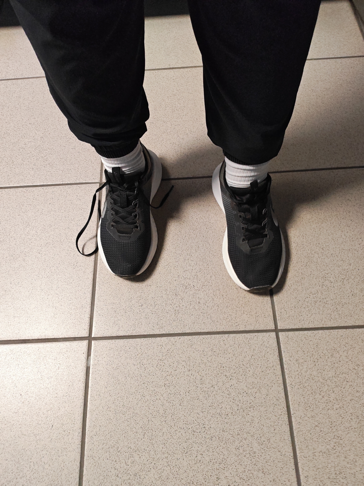 The image shows a close-up of a person's feet wearing black athletic shoes and white socks, standing on a tiled floor. The shoes appear to be Nike brand, with the distinctive swoosh logo visible on the side of the right shoe. The laces on the left shoe are untied, with one lace trailing onto the floor. The person is wearing black pants that are gathered at the ankles. The tiled floor is light beige with a speckled texture, divided by dark grout lines. The lighting suggests an indoor setting, possibly a hallway or public space, with shadows cast on the floor, indicating artificial light. There is no visible text in the image. The overall impression is casual and everyday. The specific location context of Rostov-on-Don, Russia, does not provide further visual cues in the image itself.