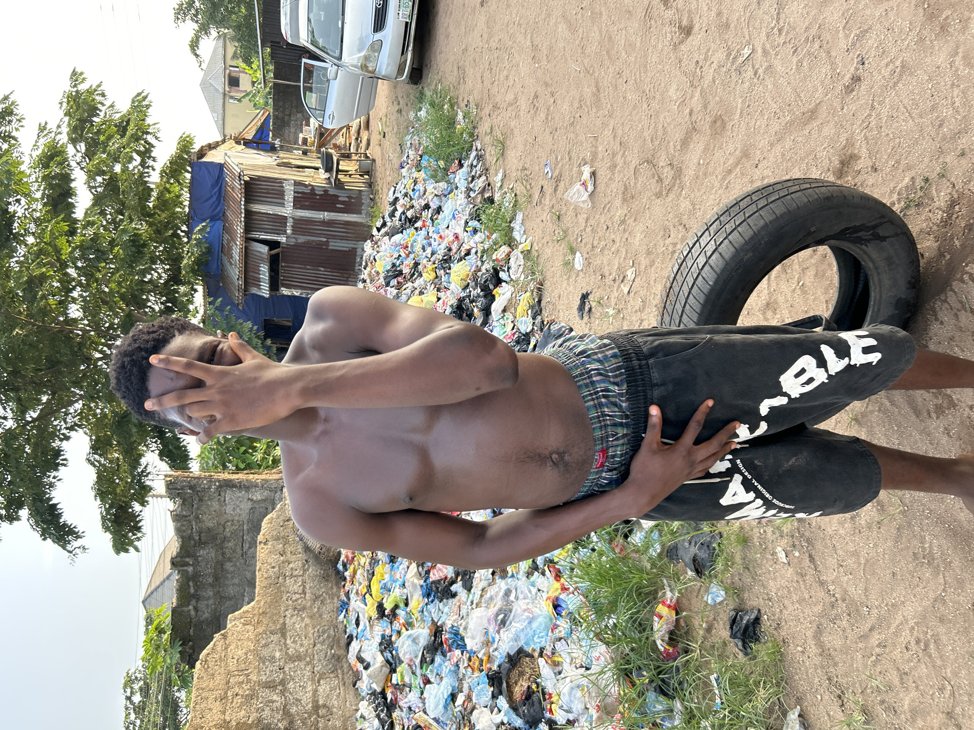 In Ihiagwa, Nigeria, a shirtless Black man stands in a littered area. He wears plaid shorts and dark-colored pants with white lettering. He partially obscures his face with his hand. Behind him, there is a large pile of plastic waste and debris. In the background are some buildings, a vehicle, and vegetation. A tire sits near him on the ground.