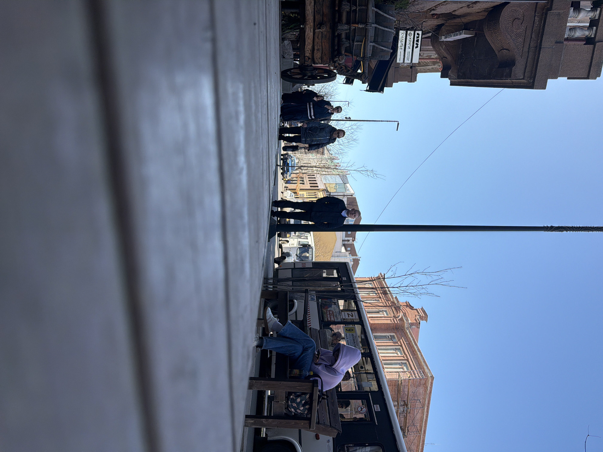 This outdoor street scene, set in Krasnoyarsk, Russia, captures a sunny day with a clear blue sky. The perspective is from a slightly low angle, looking across a street or pedestrian area lined with buildings and people.
In the immediate foreground, a person is seated on a wooden bench, wearing a light purple hoodie, blue jeans, and white sneakers, engaged with their phone. A patterned backpack rests near their feet. Adjacent to the bench is a dark-colored bus or tram, featuring numbers "2" and "9" and partially visible Cyrillic text, suggesting "AUTORU..." and "...HA", likely indicating a bus stop. To the right, a multi-story brick building with ornate architectural details and numerous windows rises, characteristic of historic European or Russian urban centers.
Further down the street on what appears to be a sidewalk, several individuals are visible. One man in a dark jacket stands closer to the foreground, while a group of three people in dark attire is positioned further back, near a large, antique-style wooden wagon with large wheels, possibly a decorative piece or historical prop. Beside this wagon, white signs display Cyrillic text: "КРУГЛИ" (Krugli - Circles), "WAM" (Vam - To you), and "СЧАСТЬЕ" (Schastye - Happiness).
The background features more buildings and bare trees, indicative of late winter or early spring. Utility poles and thin power lines stretch across the expanse of the bright, cloudless sky. The bright sunlight and absence of clouds suggest it is daytime, likely mid-morning or afternoon. The overall atmosphere is calm, with people going about their daily routines in an urban environment.