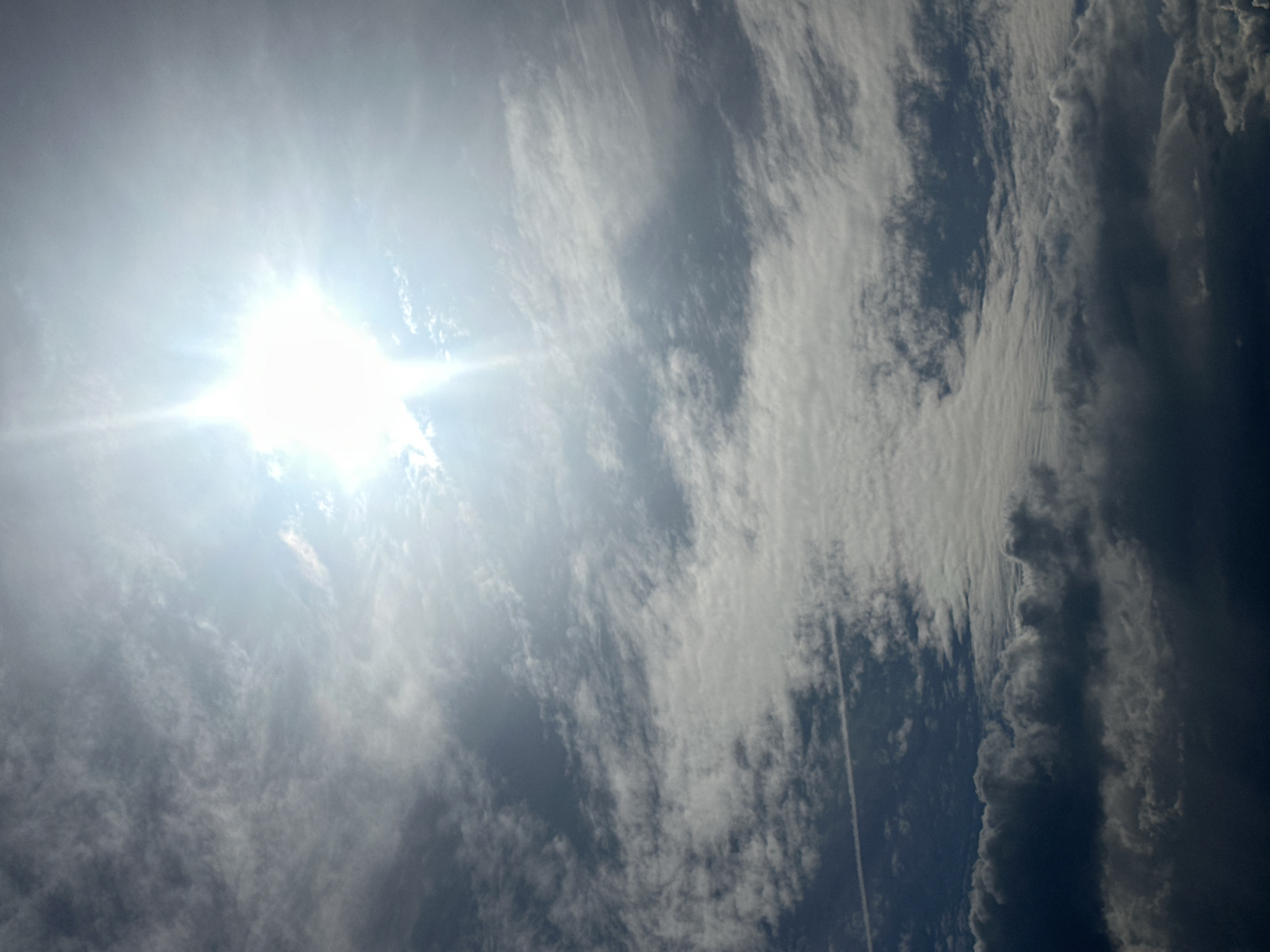 A bright sun flares in the upper right quadrant of the image, its light diffused by scattered clouds. The clouds range in color from a bright white, where they are illuminated by the sun, to dark gray and almost black at the bottom of the frame, suggesting a complex atmospheric condition. Fine, wispy cirrus clouds are visible in the mid-ground, while larger, more ominous-looking cumulus clouds are present closer to the bottom. A faint contrail from an aircraft is visible as a thin white line stretching horizontally across the left side of the image. The scene depicts a sky with a dynamic interplay of light and shadow due to the cloud cover.