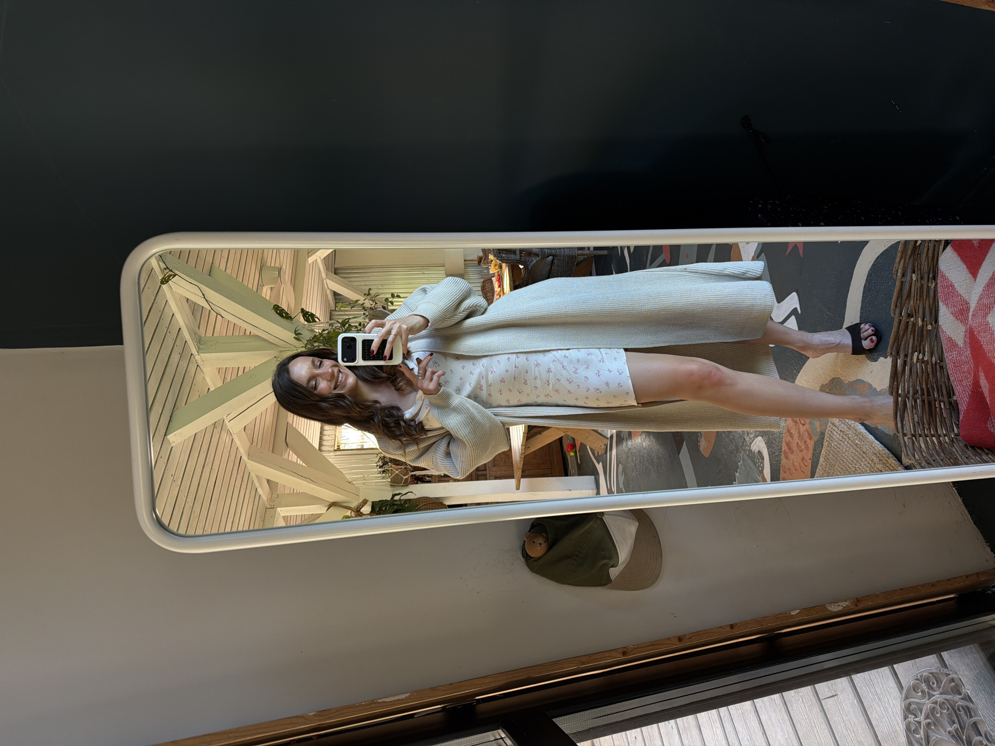 A young woman stands in front of a rectangular mirror, taking a selfie with her smartphone. She is wearing a light-colored floral dress and a long, open cardigan. The mirror reflects a scene including a sloped wooden structure, plants, and a patterned floor or rug. A hat with a small toy on top sits on a shelf or ledge behind the mirror. The area surrounding the mirror appears to be part of an interior space with a dark-colored wall on one side and a lighter-colored wall below. A window is visible at the bottom of the image. The location is not identifiable as Prudki, Russia from the image alone.