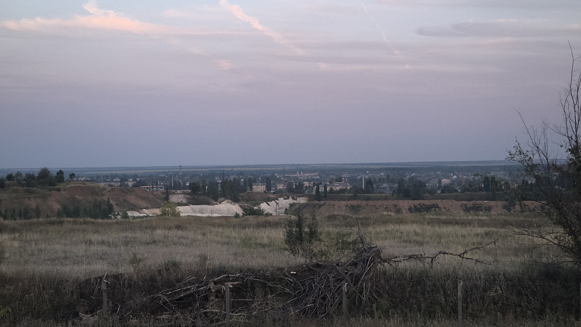 The image depicts a rural landscape at dusk, with a town or village visible in the distance. The foreground is dominated by dry, overgrown grass and scattered branches, suggesting a neglected or natural area. Beyond this, there are undulating hills with exposed soil, some of which appear to be man-made excavations or quarries, characterized by pale, sandy deposits.
Further in the background, the settlement presents a collection of buildings, including houses and industrial structures, punctuated by trees. The sky is a gradient of soft purples and blues, with faint wisps of pink clouds, indicating the transition from day to night. The overall impression is one of quietude and a somewhat desolate, yet picturesque, environment.