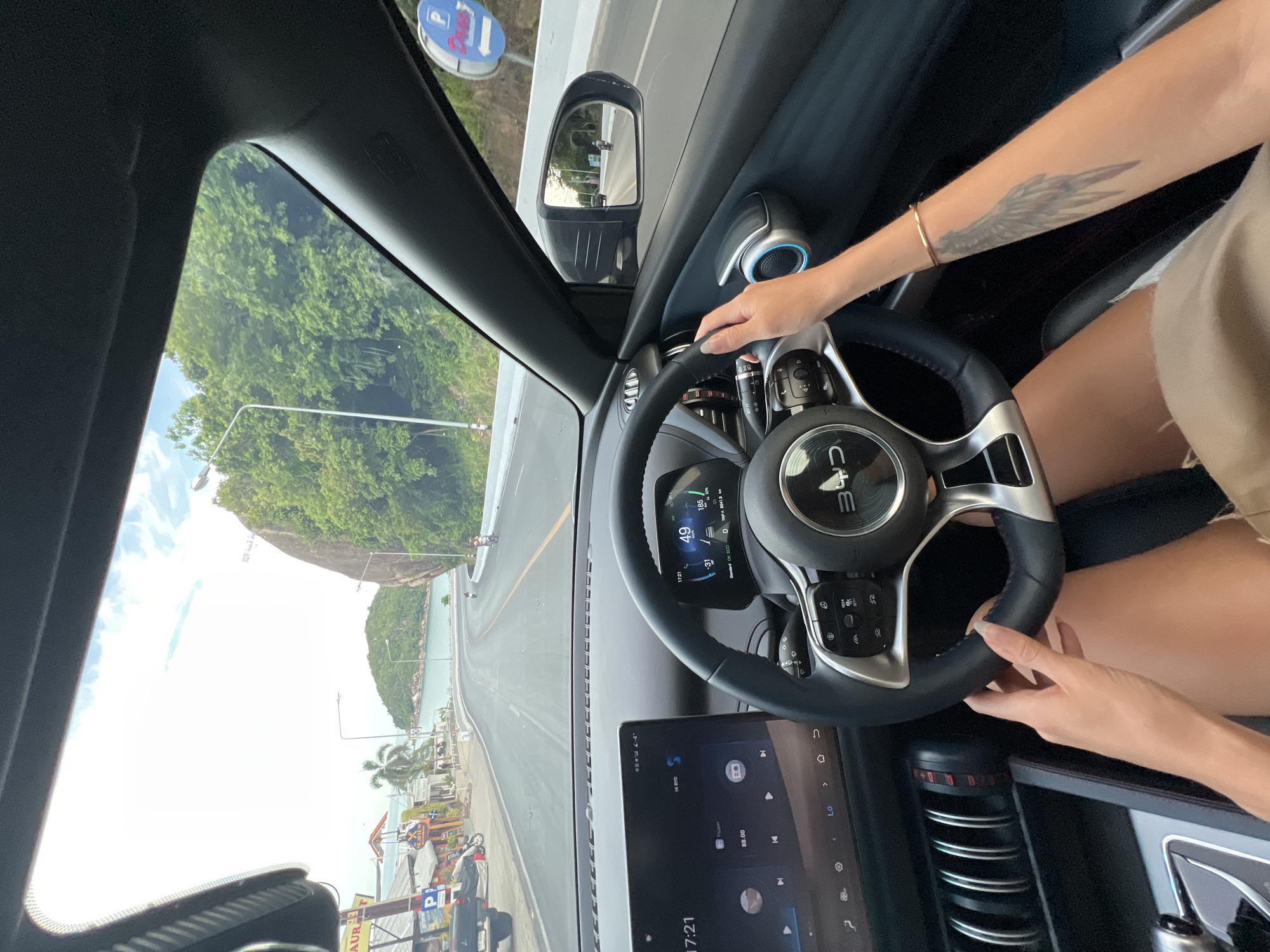The image is a first-person perspective from inside a car, driving on a coastal road. The car's interior is visible, including the dashboard with a large touchscreen display and a digital instrument cluster showing the speed as 49 km/h. The steering wheel is branded with "BYD". The driver's hands are on the steering wheel, and their legs are visible in the lower part of the frame.
Outside the car, the road curves to the right, bordered by a lush green hillside on the right and a row of shops and buildings on the left. The shops appear to be along a beach or waterfront area, with palm trees visible. In the distance, there is a body of water and islands, suggesting a tropical location. The sky is bright with some clouds. A blue road sign with a white 'P' symbol and an arrow is visible on the right side of the road. A motorcycle is seen in the side mirror, also traveling on the road. The scene suggests a sunny day, likely in Thailand, given the presence of the shops and tropical landscape. Based on the context provided about Baan Chaweng Noi, Thailand, the scene is likely depicting a drive along the coast in that area.