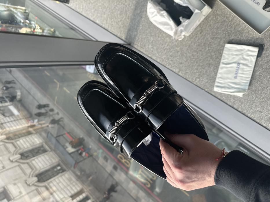 A pair of new, black, patent leather loafers with silver-tone chain-and-bar hardware is held by a human left hand in the foreground. The hand wears a dark sleeve cuff and a red string bracelet with small gold beads. The scene is viewed from an elevated position, looking down onto a street in London, United Kingdom, through a large window. On a light grey surface to the right, an open shoe box with white tissue paper and a white fabric dust bag marked "BURBERRY" are present. The exterior scene features a classic stone building with arched windows and multiple stories across the street. Below, a paved street with a distinct red line marking along the curb (indicative of a London red route or bus lane) is visible. Several blurred pedestrians are walking on the pavement, and a black bus stop shelter with a red roof, containing two seated individuals, is present. A partial street sign with the word "STUDIO" is also visible on the pavement. The overall architecture and street markings are consistent with central London.
