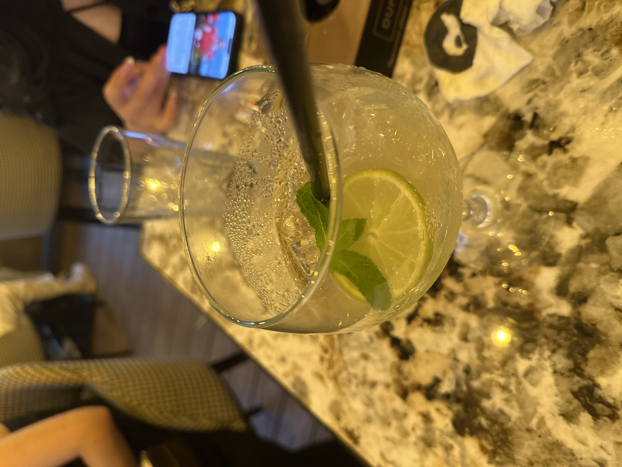 The image depicts an indoor scene at a restaurant or bar, specifically identified as Durty Kitchen & Cocktails in Sofia, Bulgaria, based on the visible "DURTY" branding on a coaster.
In the foreground, a large, bulbous goblet glass occupies the center-left, filled with a clear, light amber liquid, ice cubes, a round slice of lime, and a sprig of mint. The glass exhibits condensation on its exterior. A dark straw is partially visible, extending into the drink. The glass rests on a polished, reflective tabletop with a mottled, multi-toned pattern resembling marble or granite, featuring shades of cream, beige, brown, and dark gray/black.
To the immediate right of the glass, a crumpled white paper napkin and a black coaster or menu cover are present. The coaster bears the white, stylized text "DURTY," confirming the location.
In the midground, a person's right hand and forearm, with manicured fingernails, are visible on the right, holding a black smartphone. The phone's screen displays colorful content, predominantly red. Behind the main drink glass, another, taller cylindrical glass is partially visible, containing clear liquid and reflecting a yellow light source. To the left, the upper arm and shoulder of a person wearing dark clothing are partially visible.
The background on the left features the upholstered back of a chair, displaying a textured, possibly geometric or checkered pattern in a muted olive-green or brownish-yellow hue. Further in the background, elements of blurred furniture and indistinct figures suggest a populated indoor environment. The scene is illuminated by warm, artificial light, casting reflections on the table and glass surfaces.