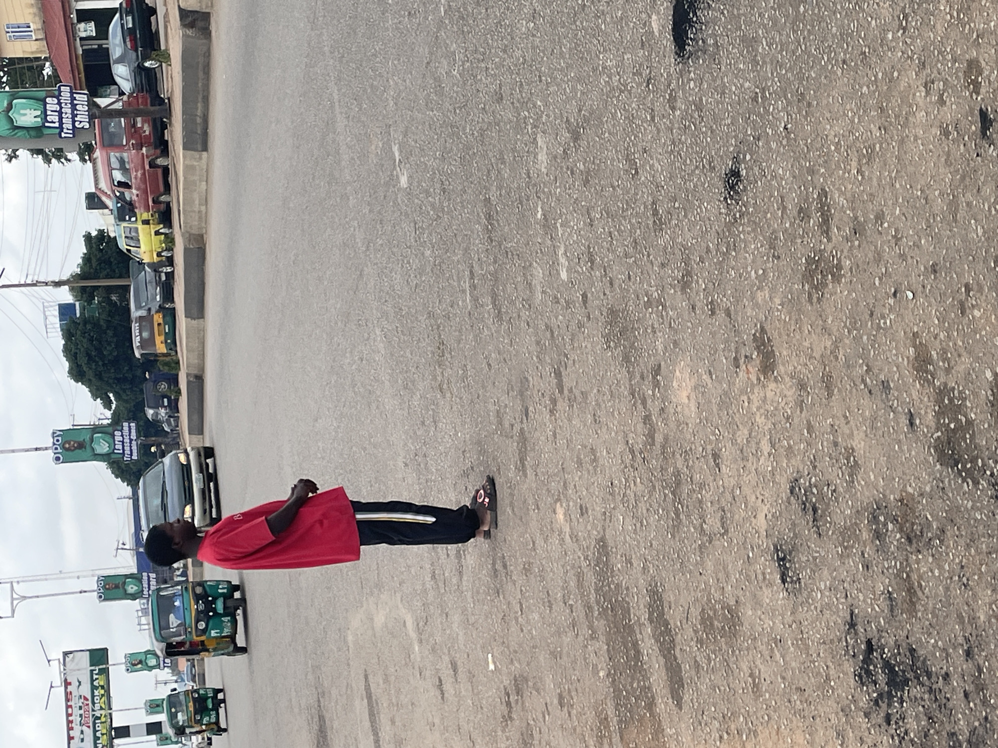 A full-shot street scene in Gura Top, Nigeria shows a young Black man in a red t-shirt and black pants with white and yellow stripes standing in the foreground facing right. He is wearing brown sandals. Behind him, a paved road has several vehicles, including three tricycles and a dark-colored van. Two large billboards are visible in the background, one advertising OPay. The road is in disrepair with visible potholes.