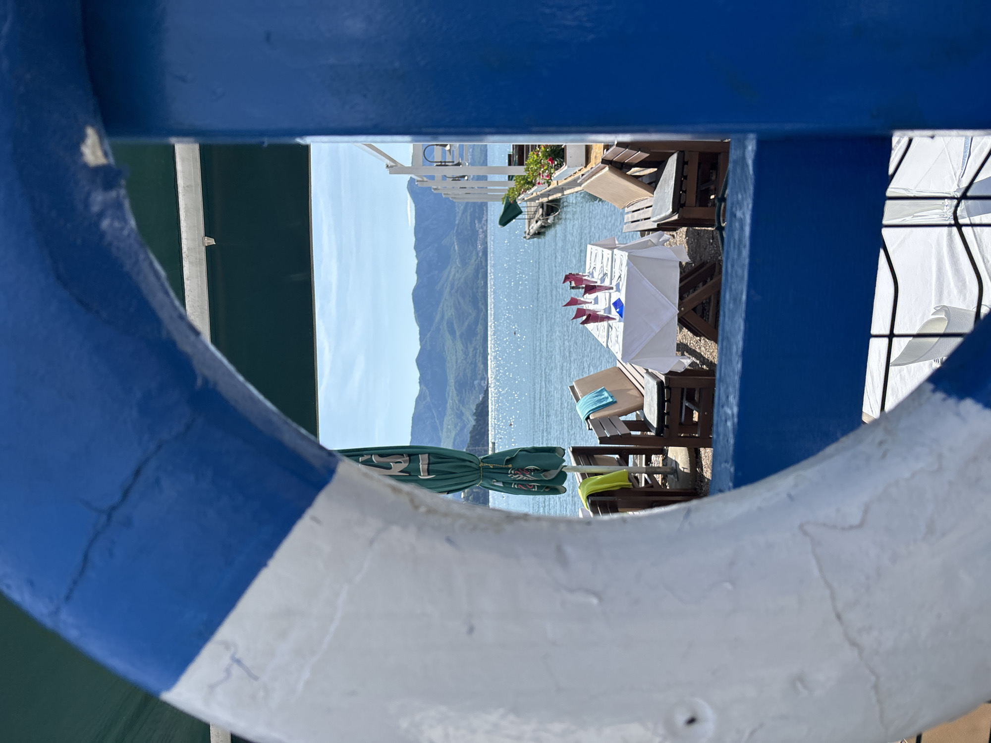 The image presents a serene waterfront scene, framed dramatically through a blue and white nautical ring, likely a lifebuoy, and parts of a blue railing or structure in the immediate foreground. The perspective is rotated 90 degrees clockwise from a standard landscape view.
In the midground, a tranquil body of water, possibly the sea or a large lake, glistens under bright sunlight, extending towards a distant shore. Majestic green mountains rise from the water, forming a scenic backdrop. Along the water's edge, several outdoor dining tables are arranged, some adorned with crisp white tablecloths. One table, centrally positioned within the frame, is neatly set with folded red napkins and small blue items, indicating readiness for diners. Nearby, wooden chairs and sun loungers are visible, with light blue and yellow towels draped over some. A closed green umbrella, bearing a white logo that vaguely resembles a deer's head, stands on the left. Portions of a white sun umbrella are also discernible on the right.
The scene depicts a peaceful, leisurely setting, likely a restaurant or resort terrace by the water. The clear blue sky and abundant sunshine suggest a pleasant day, possibly late morning or early afternoon. There are no people visible, enhancing the sense of calm and quiet anticipation. The combination of mountains, water, and dining facilities aligns well with a coastal vacation destination like Budva, Montenegro.