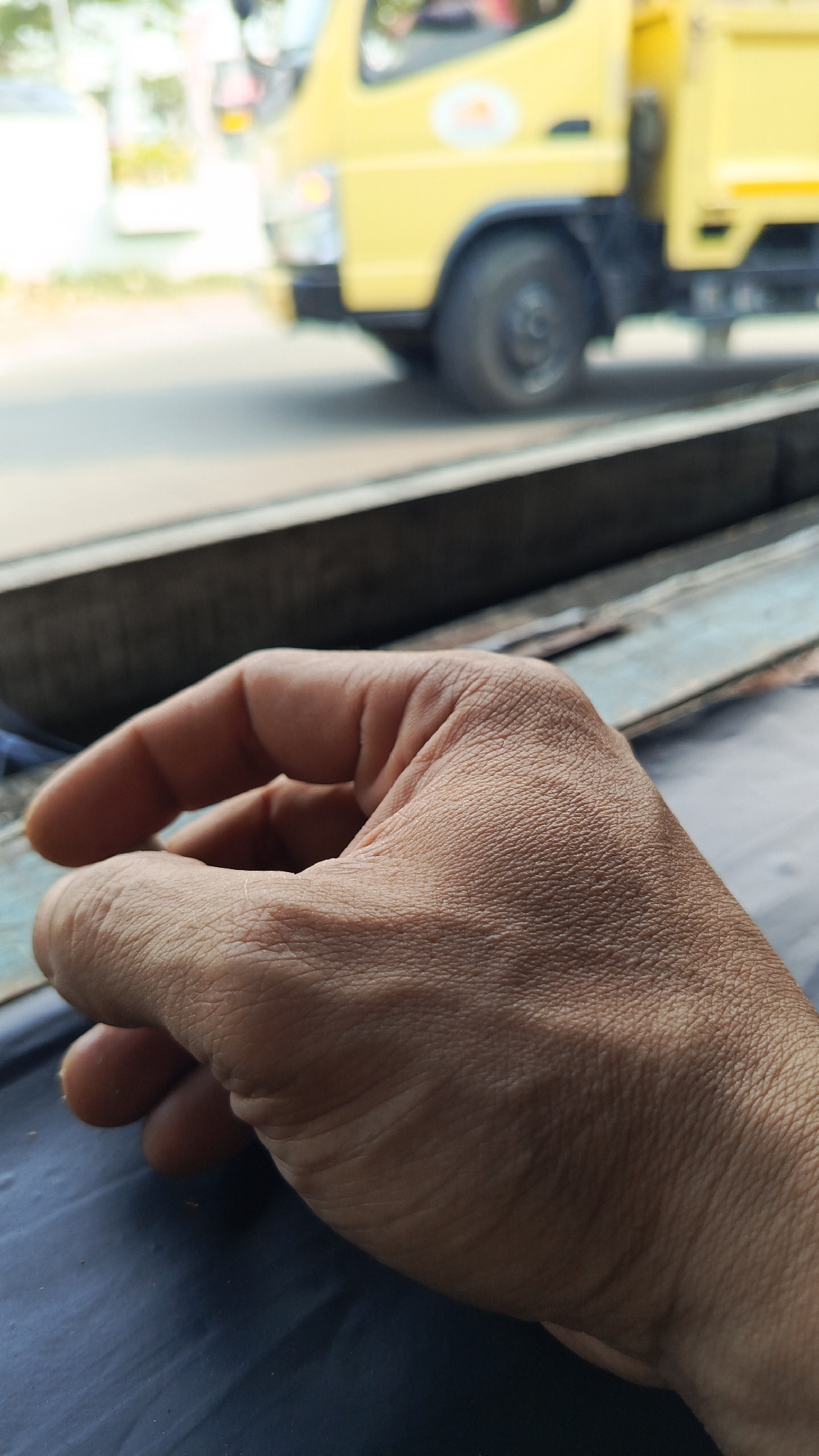 A close-up shot captures a human hand resting on a dark blue fabric surface, with a blurred yellow truck visible in the background, suggesting an outdoor urban environment. The hand, with visible wrinkles and veins, is positioned in the lower-right quadrant of the image, its fingers slightly curled. The truck, a common sight in Tangerang, Indonesia, appears to be moving or parked along a road, with its front portion and wheel partially in view. The lighting suggests it's daytime, with sunlight casting shadows. The overall scene depicts a moment of pause amidst the hustle and bustle of city life.