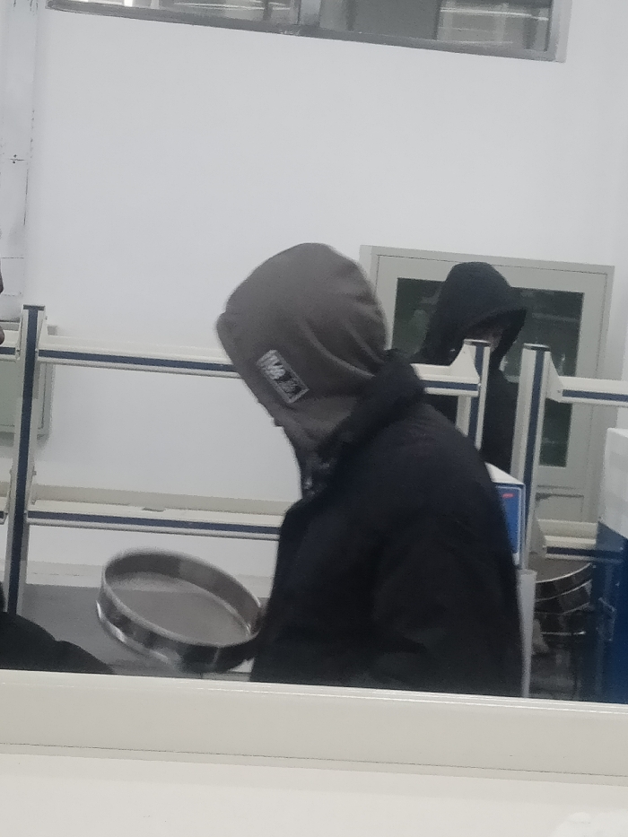 Two people in hoodies stand in a white room. One person, wearing a dark gray hoodie, looks down at a round, flat object being held up. The second person, hooded in black, stands further back. A white shelf and a metal cabinet are in the background. A partially visible desk or counter is at the bottom.