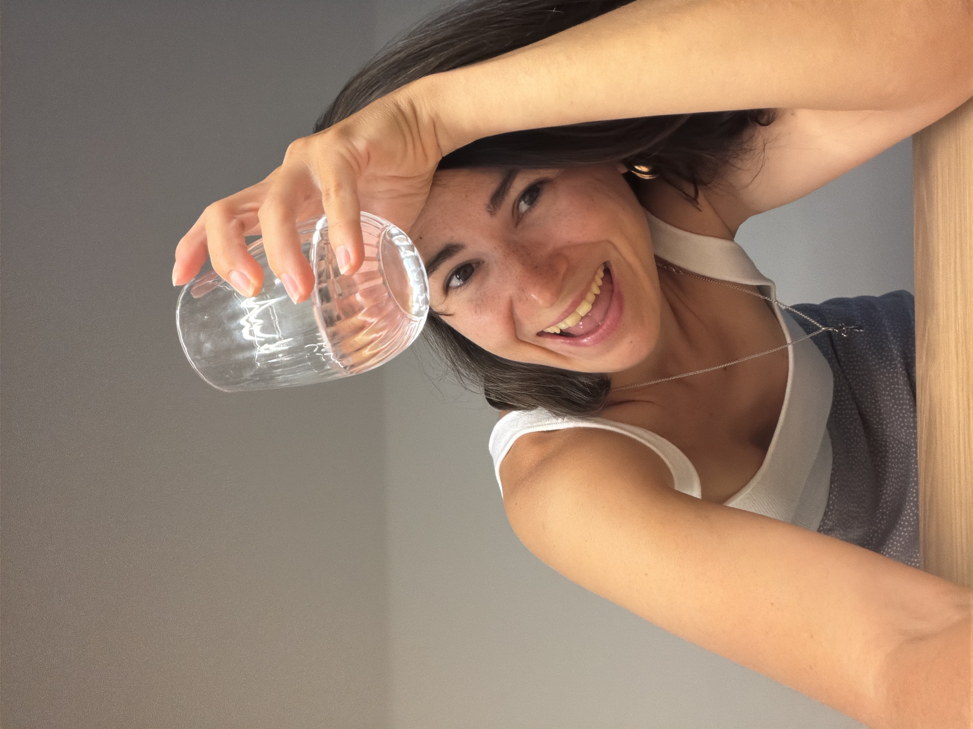 A woman is holding an empty, clear glass. She is smiling at the camera with her tongue slightly out. She has dark hair, fair skin, and freckles. She is wearing a white tank top and a silver necklace. The background is a light gray wall. A wooden structure is on the right side. The location is unknown and not necessarily in Budva, Montenegro.