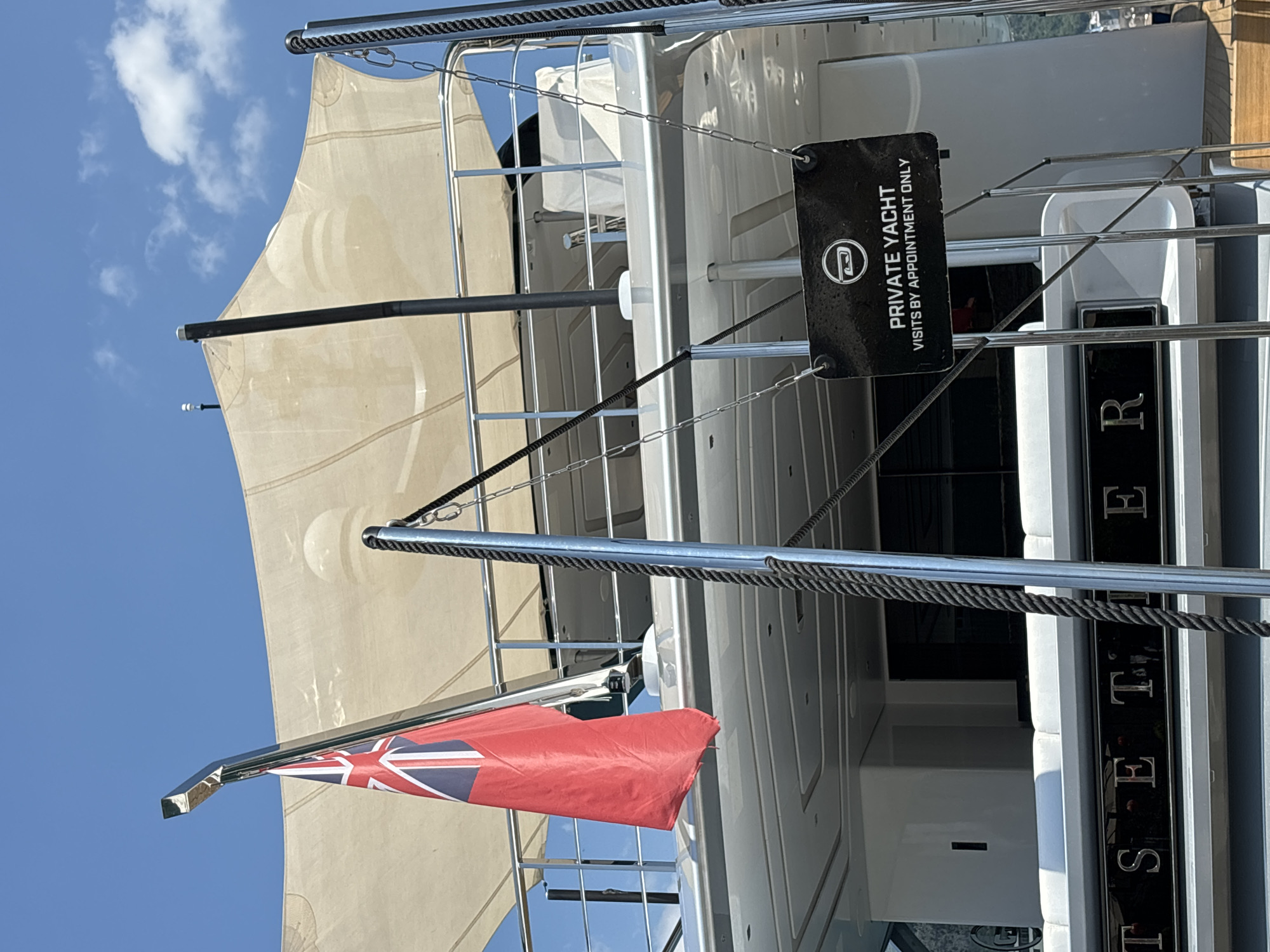 This image captures a view of a private yacht docked in Budva, Montenegro. The yacht features a red British ensign flag prominently displayed, along with a white canopy providing shade. A black sign is hung by chains, reading "PRIVATE YACHT VISITS BY APPOINTMENT ONLY" and featuring a circular logo with an abstract design. The name of the yacht, "TSETSE," is visible in silver lettering on the hull. The scene is set against a clear blue sky with a few scattered clouds, and a glimpse of the coastline with buildings can be seen in the background. The overall impression is one of luxury and exclusivity.