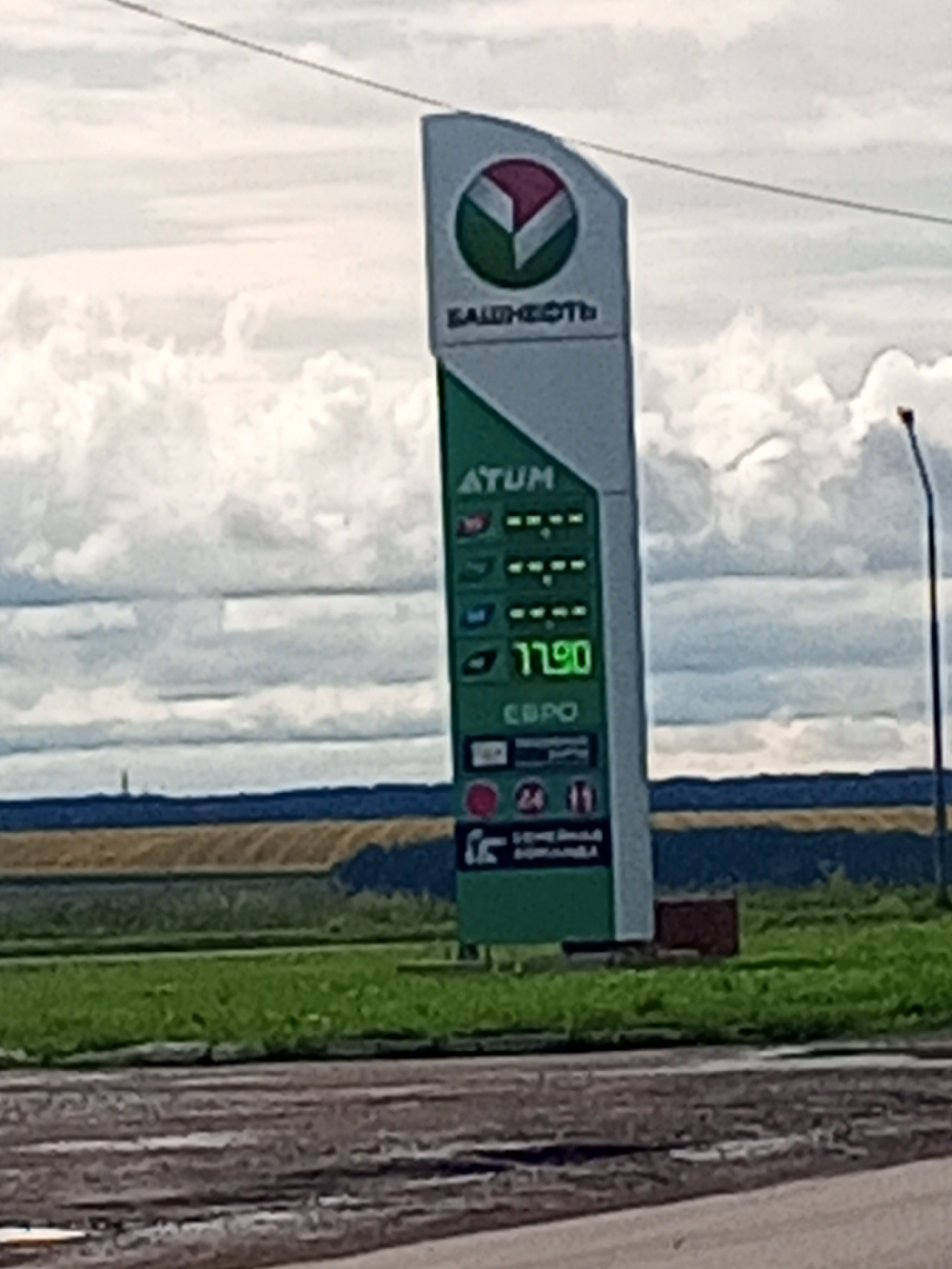 The image shows a gas station price board in the city of Kushulevo, Russia. The price board displays prices for different types of fuel, with the price for one type being 77.90. The gas station has a distinctive green and white color scheme. The sky is cloudy, and there is a field and a road visible in the background. The overall scene is that of a typical gas station on a cloudy day in a rural or semi-rural setting in Russia.