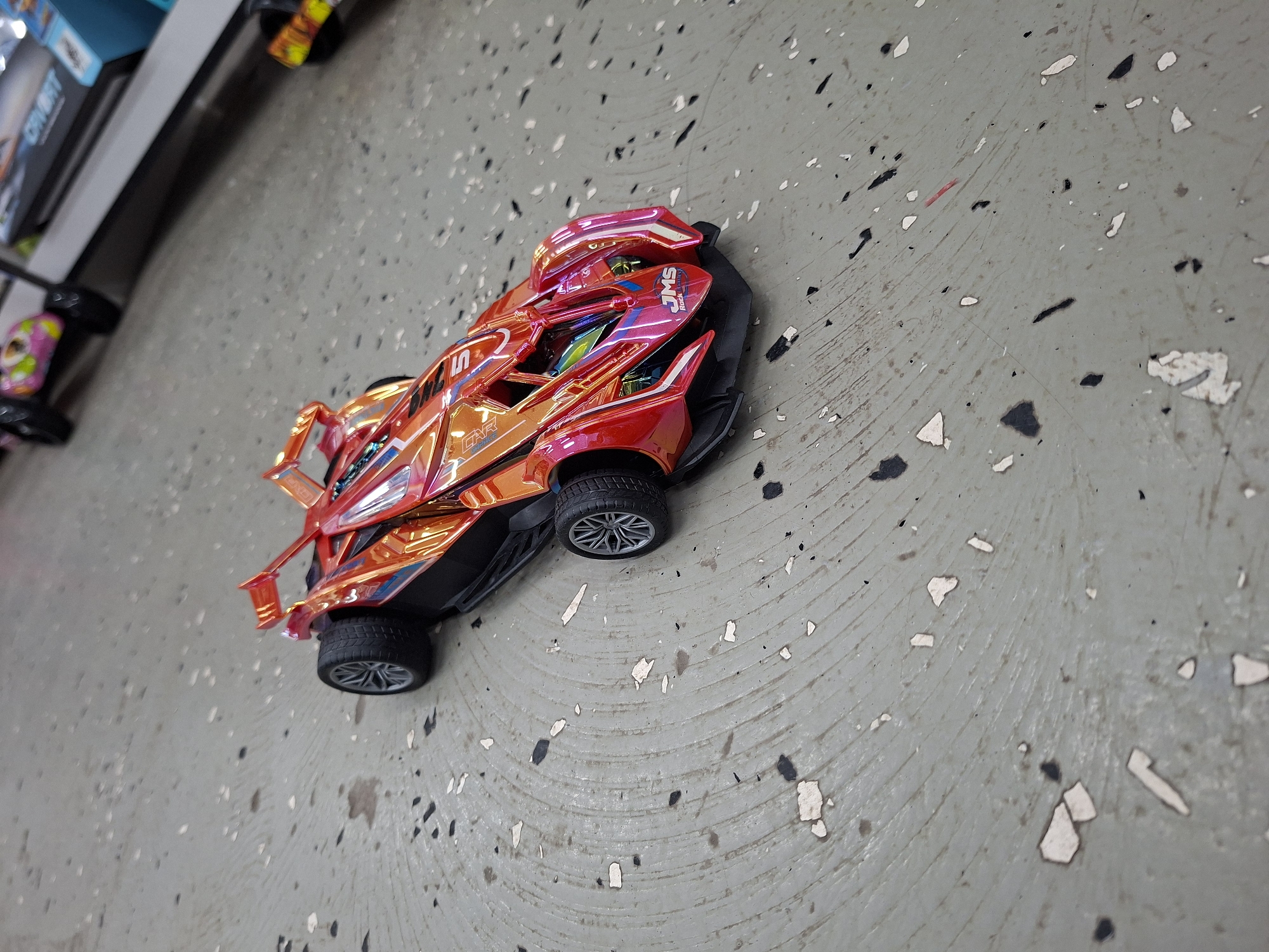 A red and orange iridescent toy race car with black tires and silver rims sits on a speckled gray floor. The car has a sleek, futuristic design with aerodynamic fins and a visible engine compartment. There is text on the car that reads "JMS Rock Energy." The floor appears to be concrete with circular markings and white and black flecks. The background is out of focus and shows shelves with what appear to be toys or merchandise. There are no people or discernible city landmarks visible. The scene does not provide any information about the city of Shira, Russia.