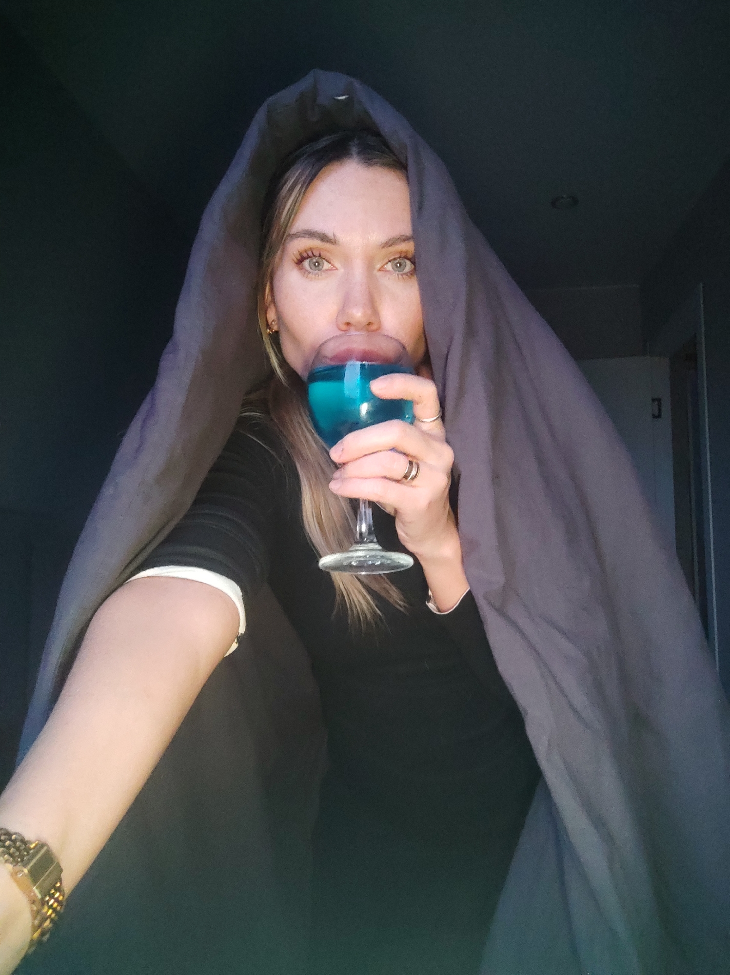 A young to middle-aged woman is depicted in a close-up, chest-up shot. She is partially shrouded by a dark grey fabric, possibly a blanket or hood, which frames her head and shoulders against a dark background. Her eyes, which appear light blue or green, gaze directly at the viewer. Her light-colored hair is visible on either side of her face. She wears a dark long-sleeved top, with a white cuff detail visible on her left wrist, alongside a gold-toned watch with a textured band.
In her right hand, adorned with two rings (one thin, one thicker with a darker center), she holds a clear stemmed wine glass filled with a bright, luminous blue liquid. Her lips are parted as she drinks from the glass. The low-light setting and indistinct background suggest an indoor scene, possibly at night or in a dimly lit room, with only subtle architectural cues like a door frame visible to the right. The overall mood is somewhat intimate and direct. No text is visible in the image. The scene takes place in Votkinsk, Russia.