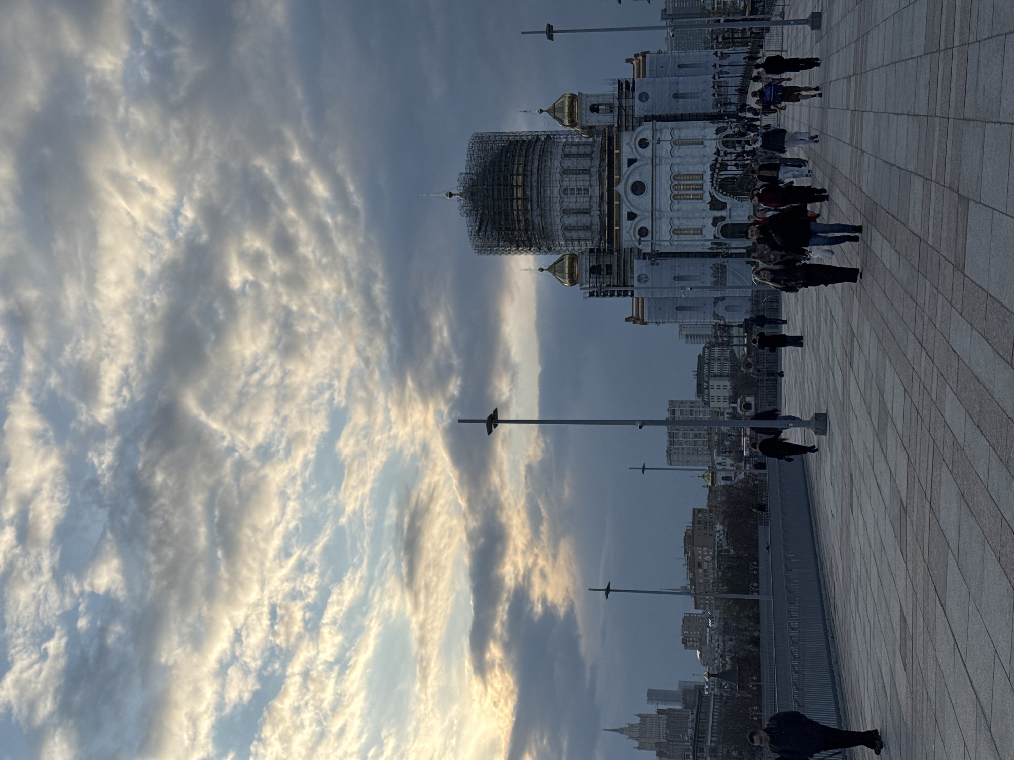 The image shows the Cathedral of Christ the Savior in Moscow, Russia. The cathedral's dome is under construction. People are walking on a wide, paved plaza in front of the cathedral. In the background, there are other buildings and a cloudy sky.