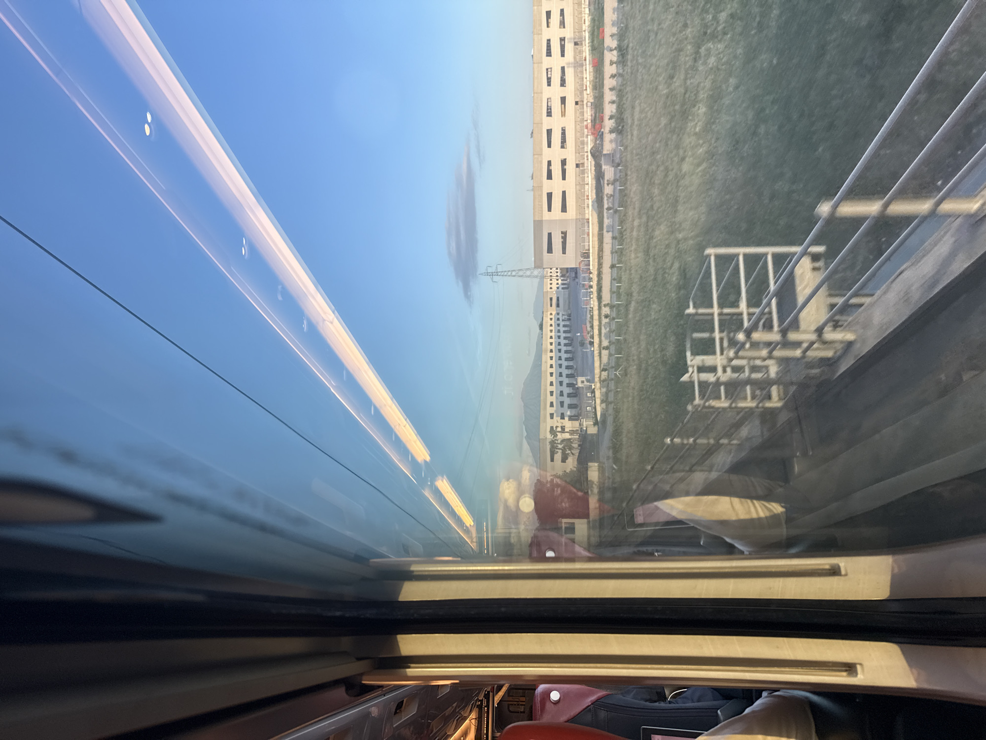 A distorted view from inside a moving train shows the landscape outside. The train's interior is partially visible in the bottom of the frame, featuring seats, luggage, and interior lighting. The window's reflection causes a mix of the interior and exterior views.
Outside the train, a building with multiple floors and regularly spaced windows dominates the view. A green field extends in front of the building, leading to a utility tower and more buildings in the distance. The sky is blue with a single, elongated cloud.
The scene is likely in Caivano, Italy, given the prompt's context.