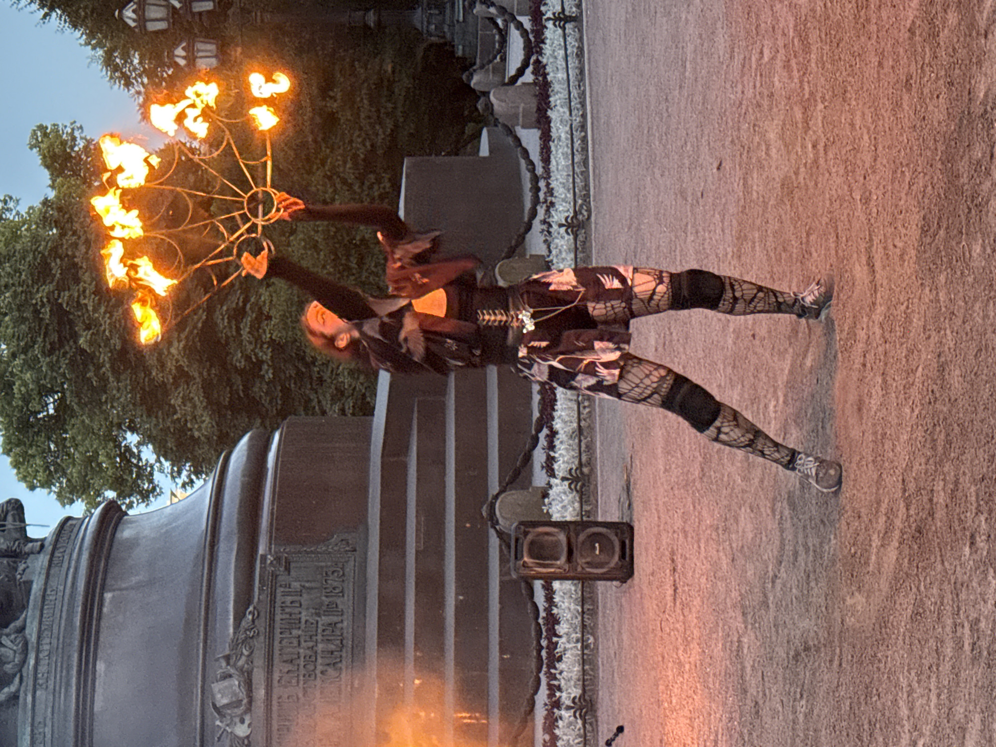 The image depicts a fire performer in front of the monument to Catherine the Great in Palace Square, Saint Petersburg, Russia. The performer, dressed in dark clothing with a patterned skirt and fishnet stockings, is holding a fan-like apparatus ablaze with multiple flames. The monument, a large bronze structure, is visible in the background, featuring steps and an inscription. A portable speaker sits on the ground to the left of the performer, and streetlights and trees are also present in the background. The scene appears to be set at dusk, with the sky displaying a soft, bluish hue.