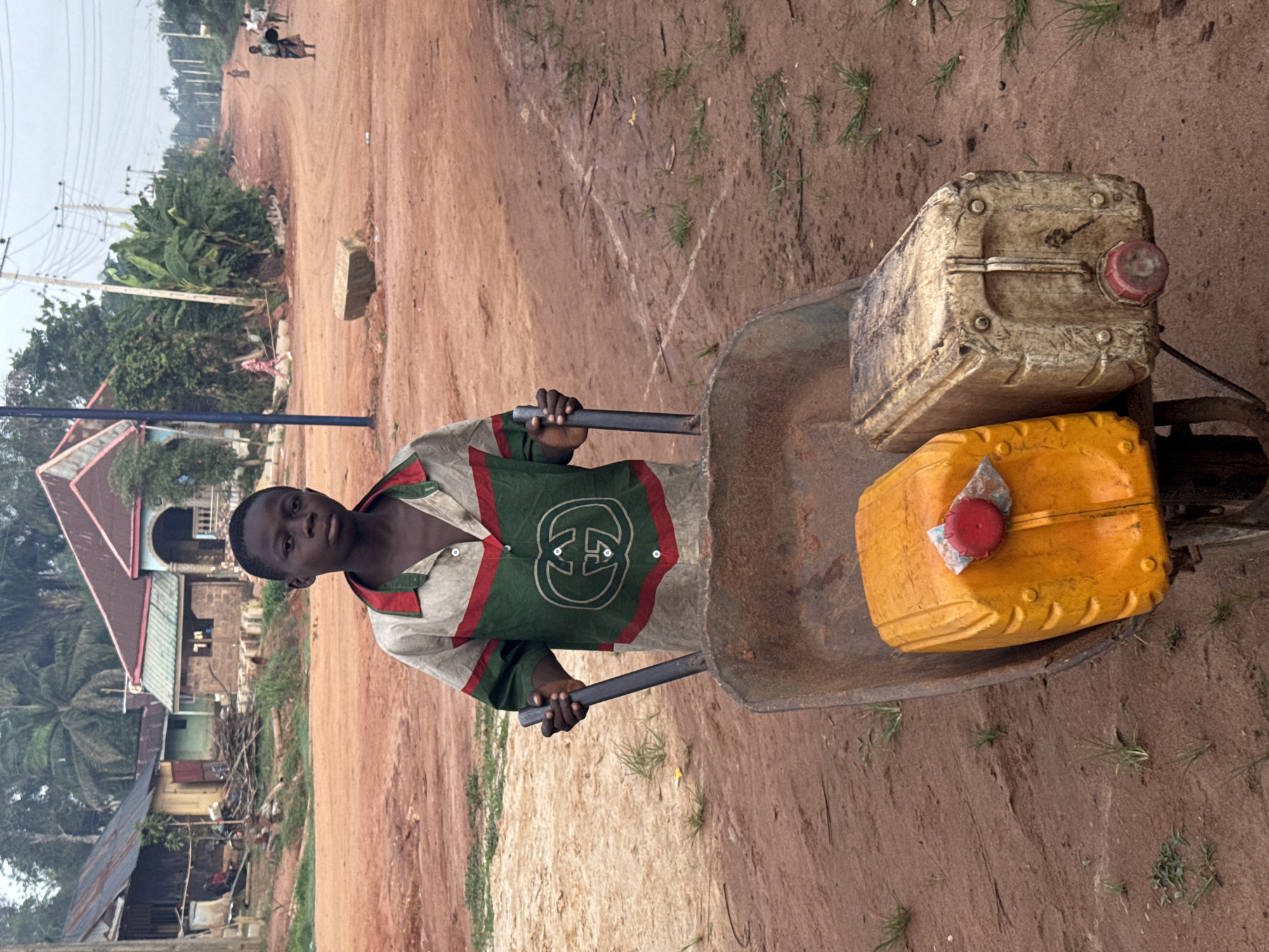 A young man stands on a dirt road in Eziudo, Nigeria, pushing a wheelbarrow containing two yellow and white jerrycans. The man is wearing a shirt with a green and red stripe pattern and a large Gucci logo. The road appears to be under construction. Buildings, trees, and electrical poles are visible in the background. Two people can be seen walking in the distance.