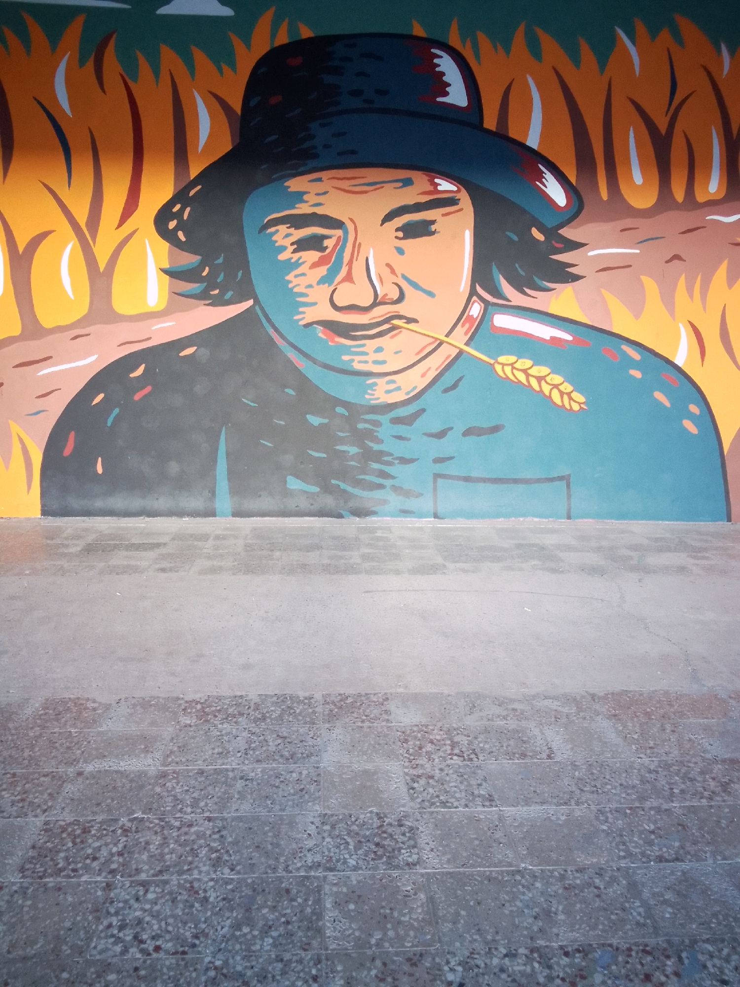 The image shows a mural painted on a wall, depicting a person wearing a wide-brimmed hat and holding a stalk of wheat in their mouth. The mural is set against a background of stylized flames or tall grass. The lower part of the image shows a concrete ground surface with a pattern of rectangular tiles. This mural is located in Tbilisi, Georgia, on the side of a building facing a pedestrian walkway. The style of the mural is flat, with bold outlines and vibrant colors, suggesting a folk art or social realist influence. The person in the mural appears to be of indeterminate gender, with a simplified, almost mask-like face. The overall impression is one of rural life and harvest.