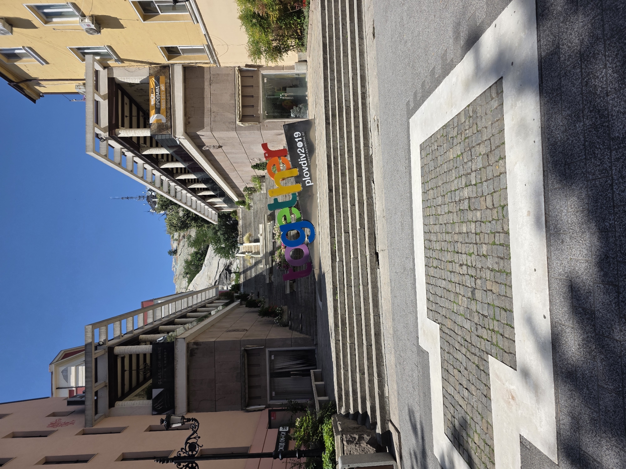 The image shows a street scene in Plovdiv, Bulgaria, specifically at the staircase leading up to the Astronomical Observatory on Sahat Tepe. In the foreground, a street is paved with asphalt and features a decorative pattern of cobblestones set within concrete borders, resembling a heart shape. A large, colorful sculpture spelling out "together" in bold, multi-colored letters is prominently displayed on a black base. Next to the sculpture, a black sign reads "Plovdiv2019 EUROPEAN CAPITAL OF CULTURE."
Flanking the staircase are two buildings. The building on the left is a light peach color with a dark lamppost in front of it. A sign above a window reads "GLASS OF WINE EST. & LOUNGE." The building on the right is yellow and has several windows. A banner on this building reads "ПРОДАВА" (For Sale) in Cyrillic script. The staircase itself is wide and made of stone, with planters containing greenery and flowers on either side. In the background, the top of a telecommunications tower is visible on a hill, consistent with the location of Sahat Tepe. The sky is clear and blue, indicating a sunny day. There are no people visible in the image.