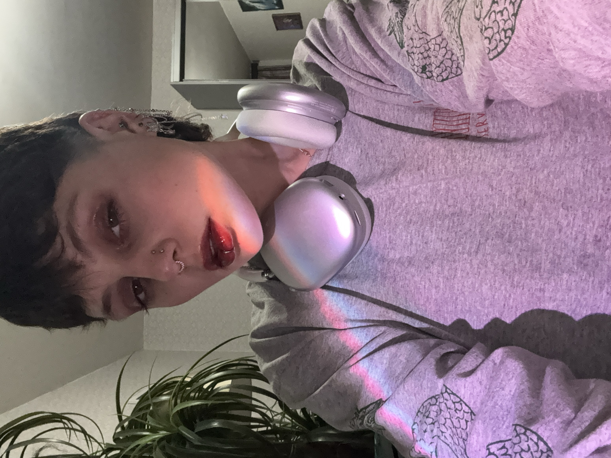 A close-up, indoor photograph shows a person with short, dark hair and multiple piercings in their nose and ear. They are wearing a grey sweatshirt with a pattern on the sleeves and silver headphones around their neck. The lighting in the room appears to have a purplish hue, with a rainbow light effect across the sweatshirt. In the background, a plant is visible, and there is a glimpse of a shelf or cabinet. The image is likely taken from a low angle, looking up at the person. No location information is available.