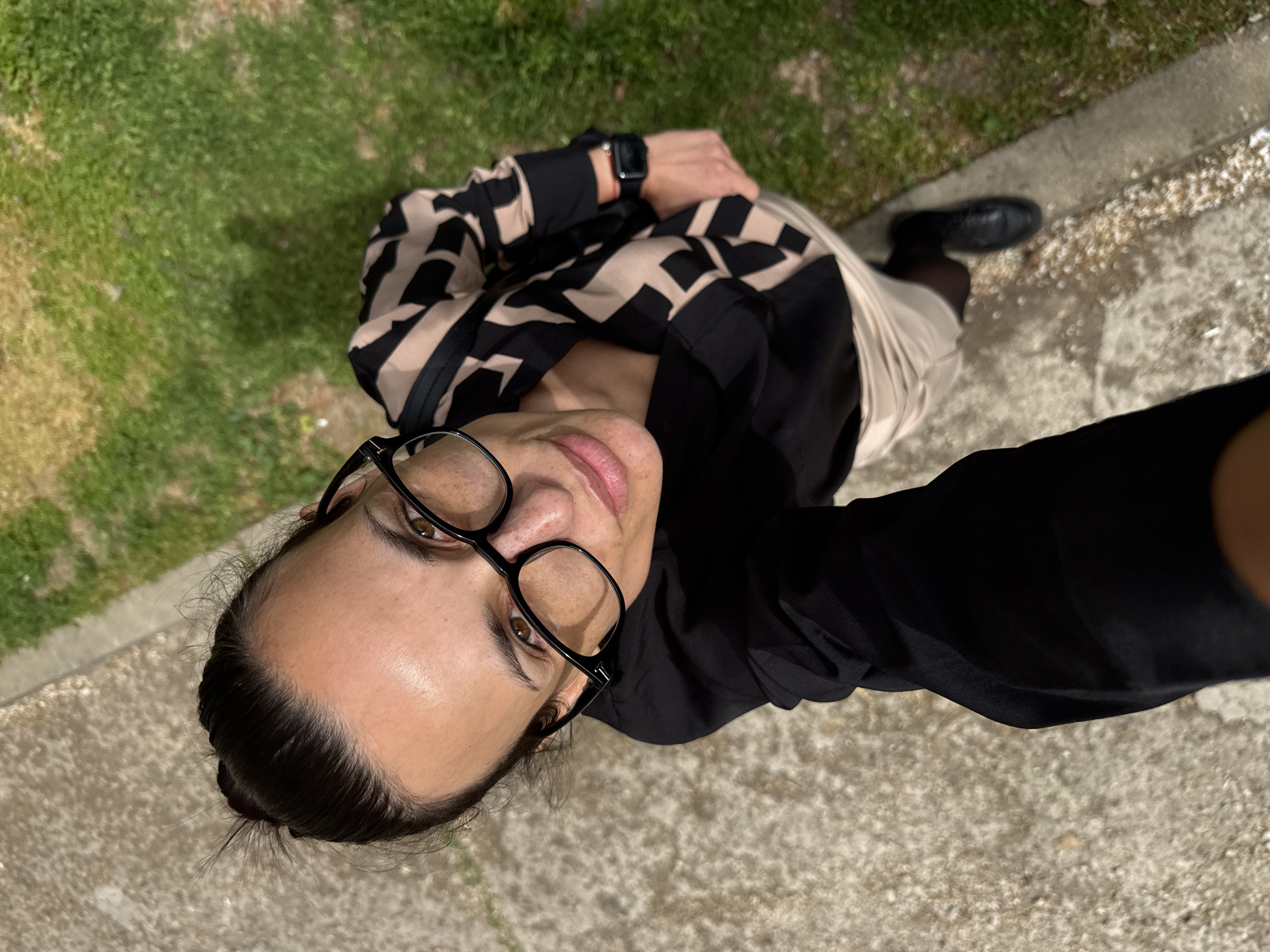 An overhead selfie captures a woman looking upwards at the camera. She has dark hair pulled back and wears black-framed glasses, with a slight, calm smile on her face. Her skin has a warm, medium tone. She is dressed in a black and beige geometric-patterned long-sleeved top worn over a black shirt, paired with a light-colored skirt or dress. A black smartwatch is visible on her left wrist. She also wears dark hosiery and black shoes.
The setting is outdoors, likely in Pleven, Bulgaria, with the woman standing on a textured, light-colored paved surface, possibly a sidewalk, with a strip of green and dry grass adjacent to it. Small, light-colored debris, possibly petals or tiny stones, are scattered on the paved ground. The lighting suggests it is daytime, with natural sunlight illuminating the scene. The overall impression is a casual outdoor moment. No visible text is present in the image.