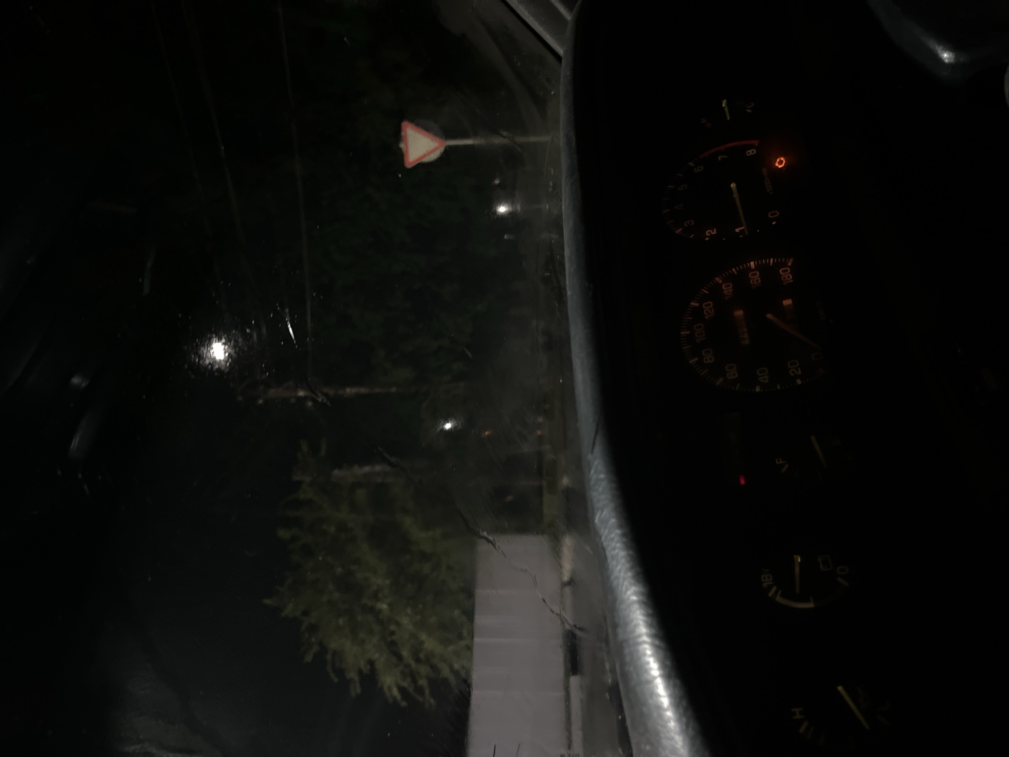 This is a nighttime, interior shot from the driver's perspective inside a vehicle, looking out through the windshield. The dashboard is partially visible in the foreground, with illuminated gauges. The car appears to be on a street or road. Outside the car, trees are visible, illuminated by streetlights or ambient light. A yield sign is positioned on the right side of the road. The windshield has streaks and appears wet, suggesting it might be raining or has recently rained. The overall ambiance is dark and atmospheric, typical of a nighttime scene.
The location of Belogorsk, Russia is not directly discernible from the visual elements in the image itself. The image depicts a generic nighttime street scene that could be found in many locations.