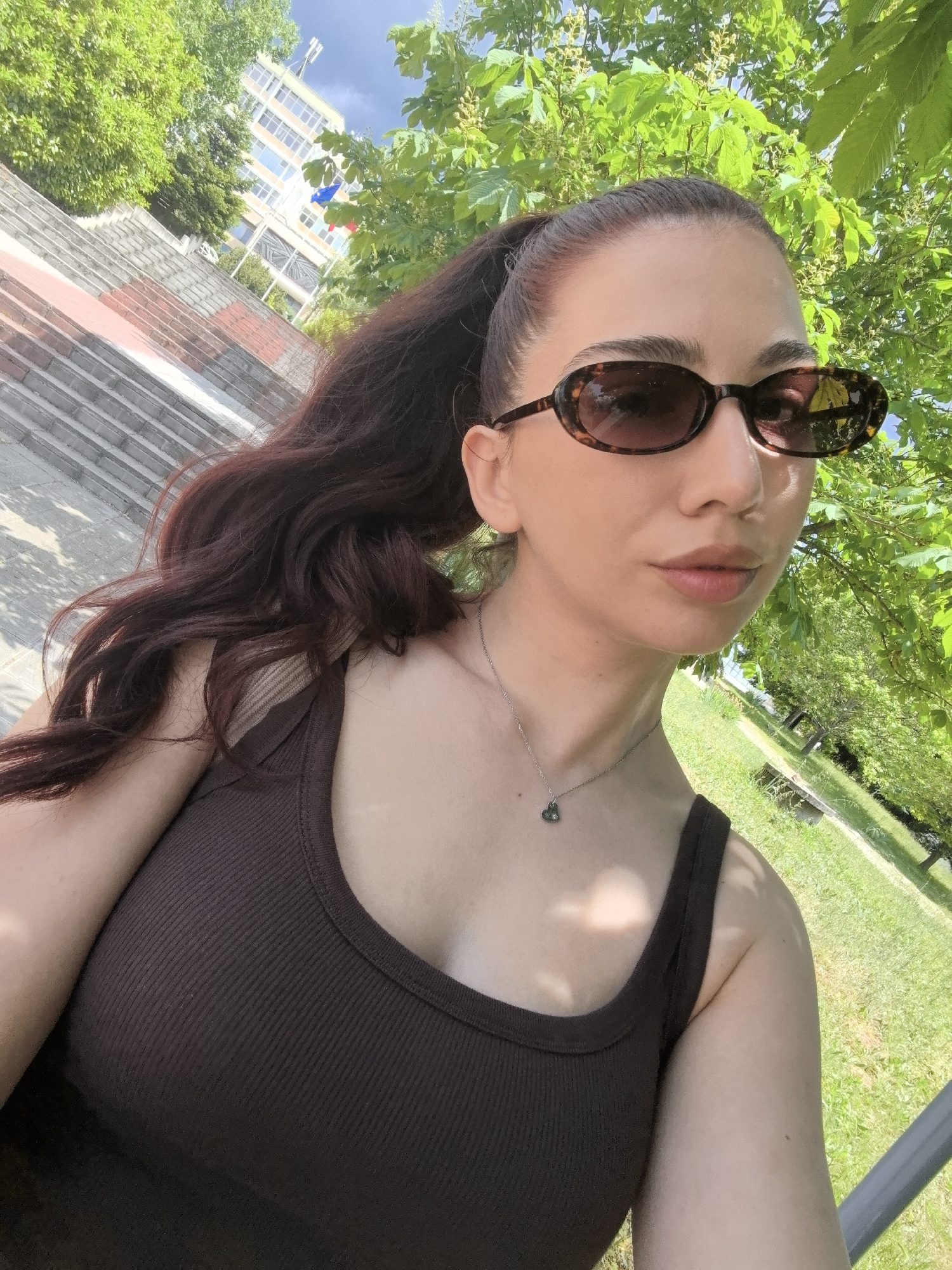 A woman with brown hair pulled back into a ponytail, wearing brown sunglasses and a brown tank top, stands in a park setting. A silver necklace with a pendant is visible. The background features green trees, a building with flags, and stone steps. The scene is in the city of Unknown, Unknown.