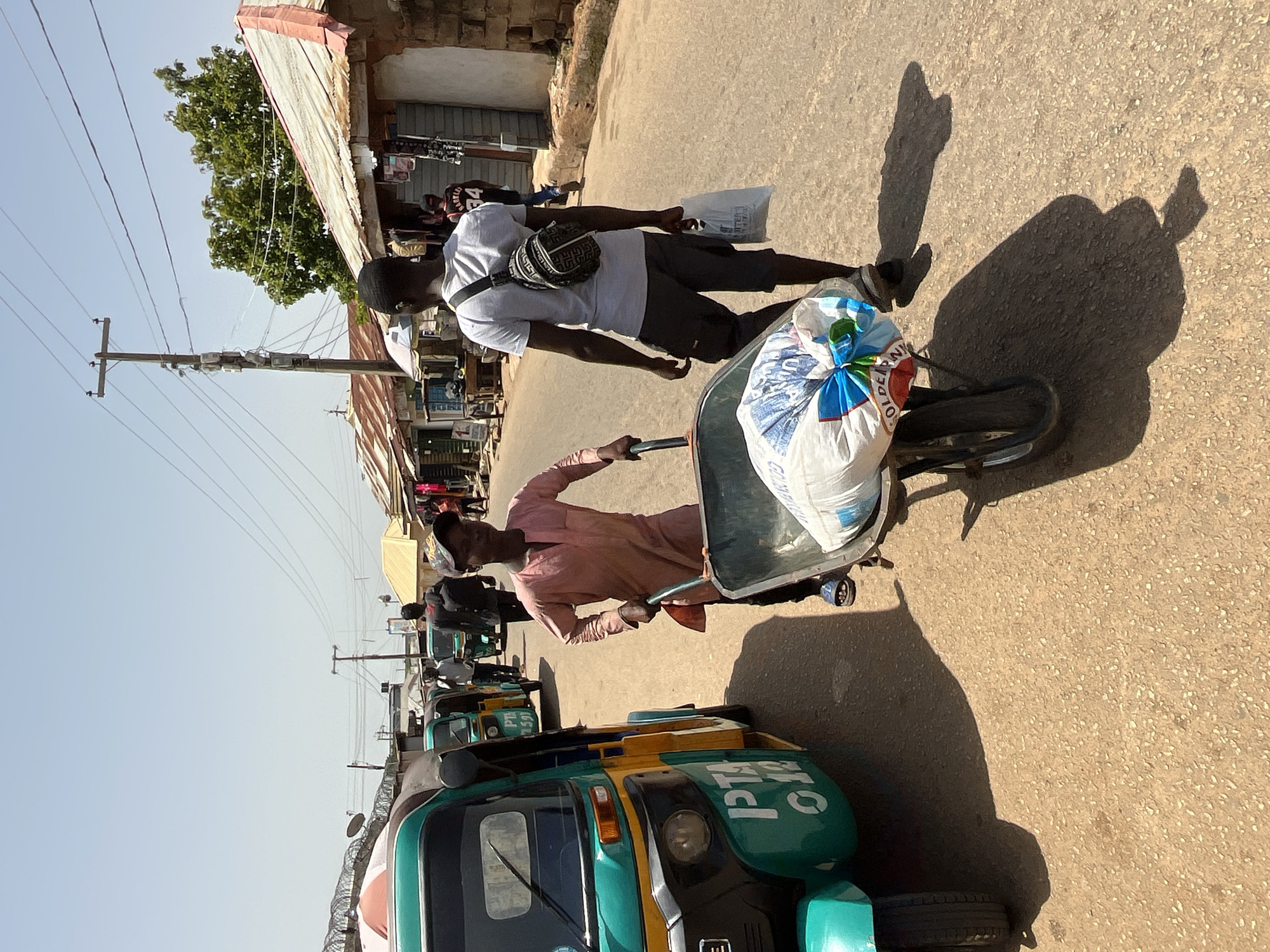 In an outdoor, dusty street scene in Unknown, Unknown, a young man wearing a pink tunic and dark pants pushes a wheelbarrow loaded with a large white sack. The sack, tied with a blue and green ribbon, appears to contain grain or a similar commodity. He is walking away from the viewer and slightly to the left. To his right, another young man, wearing a grey t-shirt and dark shorts, walks in the same direction, carrying a white plastic bag in his right hand and a small black crossbody bag over his left shoulder.
In the foreground and to the left, a green and yellow three-wheeled auto-rickshaw is parked. Several other auto-rickshaws are visible parked further down the street. The street is lined with small, weathered market stalls with corrugated metal roofs. Power lines and a utility pole are present overhead. The sky is clear and bright. The overall impression is of a busy marketplace.