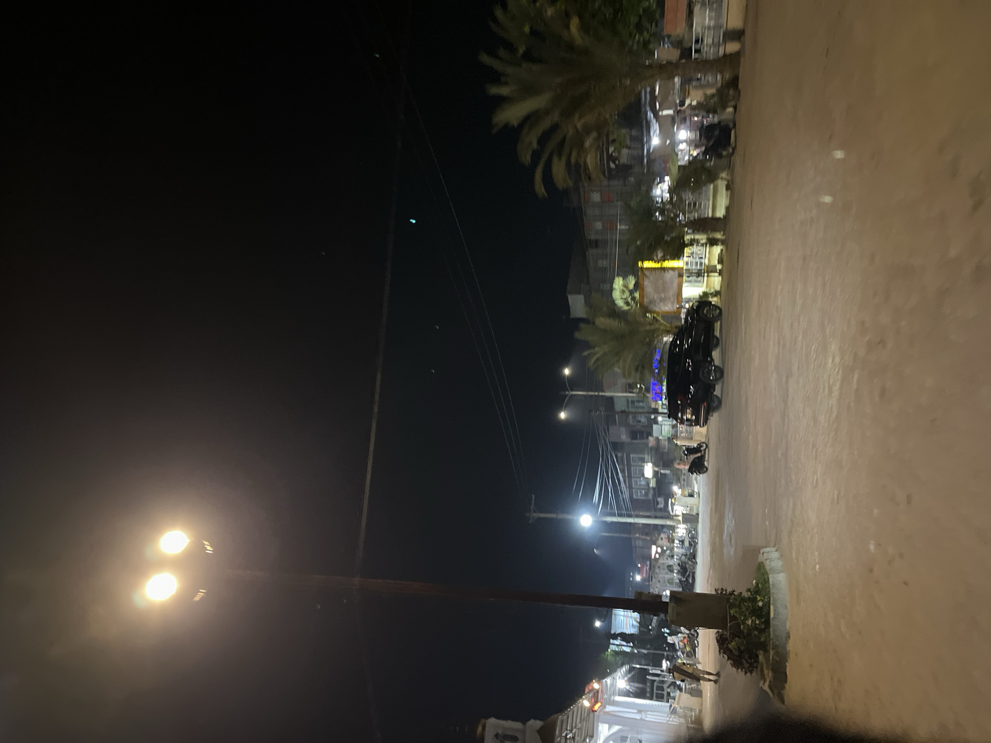 This media file captures a nighttime scene in Bundar, Indonesia. The image depicts a street or walkway lined with buildings and palm trees. Several streetlights are visible, illuminating the area with a warm glow. A black SUV is parked on the right side of the road, and what appears to be a motorcycle is parked behind it. Further down the street, more buildings can be seen, some with illuminated windows. There are also a few individuals visible, walking or standing near the buildings. The ground appears to be paved, with some areas covered in sand or dirt. The overall atmosphere is that of a quiet evening in a developing urban or semi-urban area. The time of day is clearly night, and there are no indications of specific weather conditions. The presence of palm trees and the style of the buildings suggest a tropical or subtropical location, consistent with Bundar, Indonesia. The lighting is a prominent feature, creating a slightly hazy and atmospheric effect.