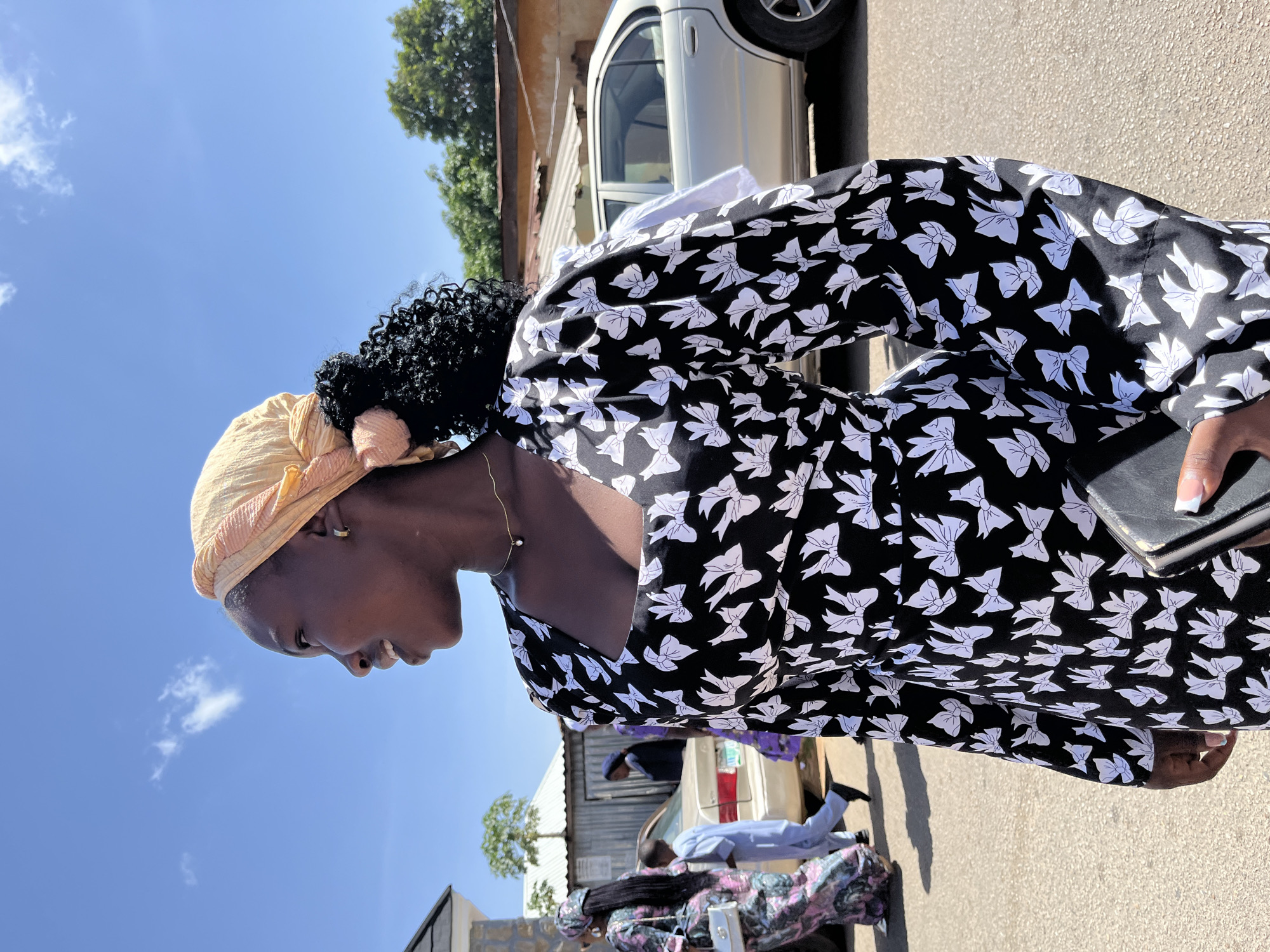 A woman with dark curly hair partly covered by a beige head wrap and wearing a black dress with a white bow pattern stands on a paved street in Jos, Nigeria. She is holding a black object in her right hand, her head slightly tilted downward. A silver car is partially visible behind her, parked on the street. Other people and another vehicle are discernible in the background near a building with a corrugated metal roof. The sky is visible in the upper left corner of the frame.