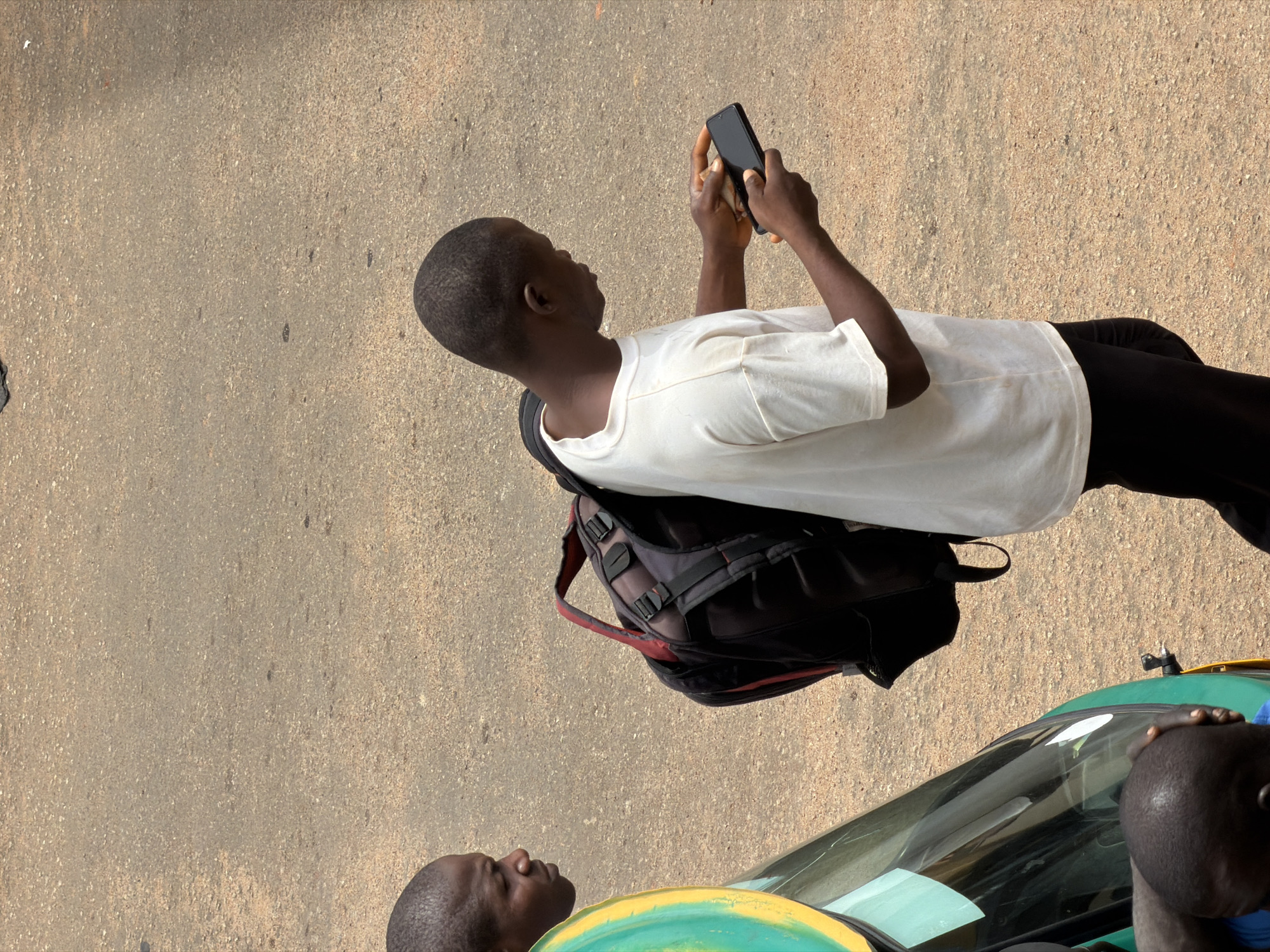 The image captures a street scene in Bukuru, Nigeria, with a focus on individuals and their surroundings. In the foreground, a person is seen from the chest up, wearing a white t-shirt and black pants, looking down at a black smartphone held in their hands. This individual has a backpack with red accents slung over their shoulders. To the left, another person's head is visible, looking towards the right. A portion of a green vehicle with a yellow stripe and a reflective surface is also visible on the far left, suggesting the presence of transportation. The background consists of a dusty, asphalt-like road surface, typical of a street environment. The lighting indicates it is daytime.