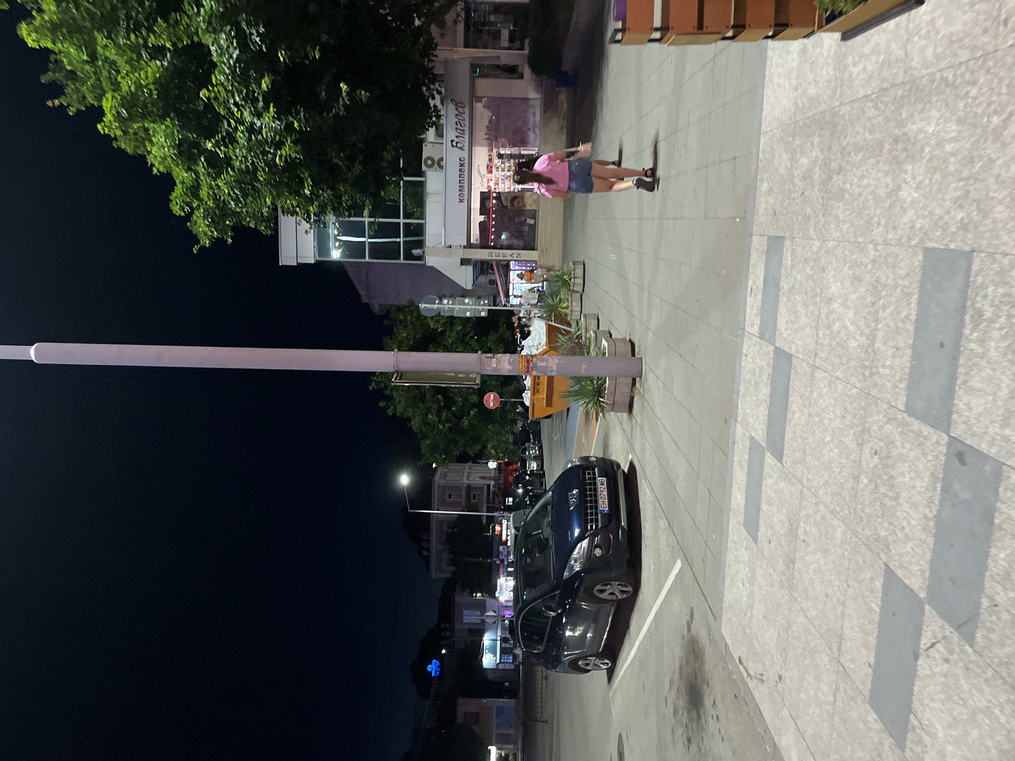 A nighttime scene depicts a street in an unknown city. In the foreground, a dark blue Peugeot SUV is parked on the right side of the frame. The car's license plate reads "EH 0792 MB". To the right of the car, a sidewalk is visible with several planters containing green foliage. Further down the sidewalk, a young woman in a pink t-shirt and denim shorts walks away from the camera, carrying a light-colored purse. She is wearing dark sandals. Behind her, more pedestrians are walking. To the right of the sidewalk, a building with a white facade and glass windows is illuminated. The building has signage in an unfamiliar script, with "КОМПЛЕКС БЛАГОЕВ" visible. A tall, pink-hued lamppost stands prominently in the middle ground, extending upwards out of frame. In the background, other buildings and streetlights are visible against the dark night sky. A large green tree with dense foliage is situated to the right of the building. The street is paved with cobblestones and concrete.