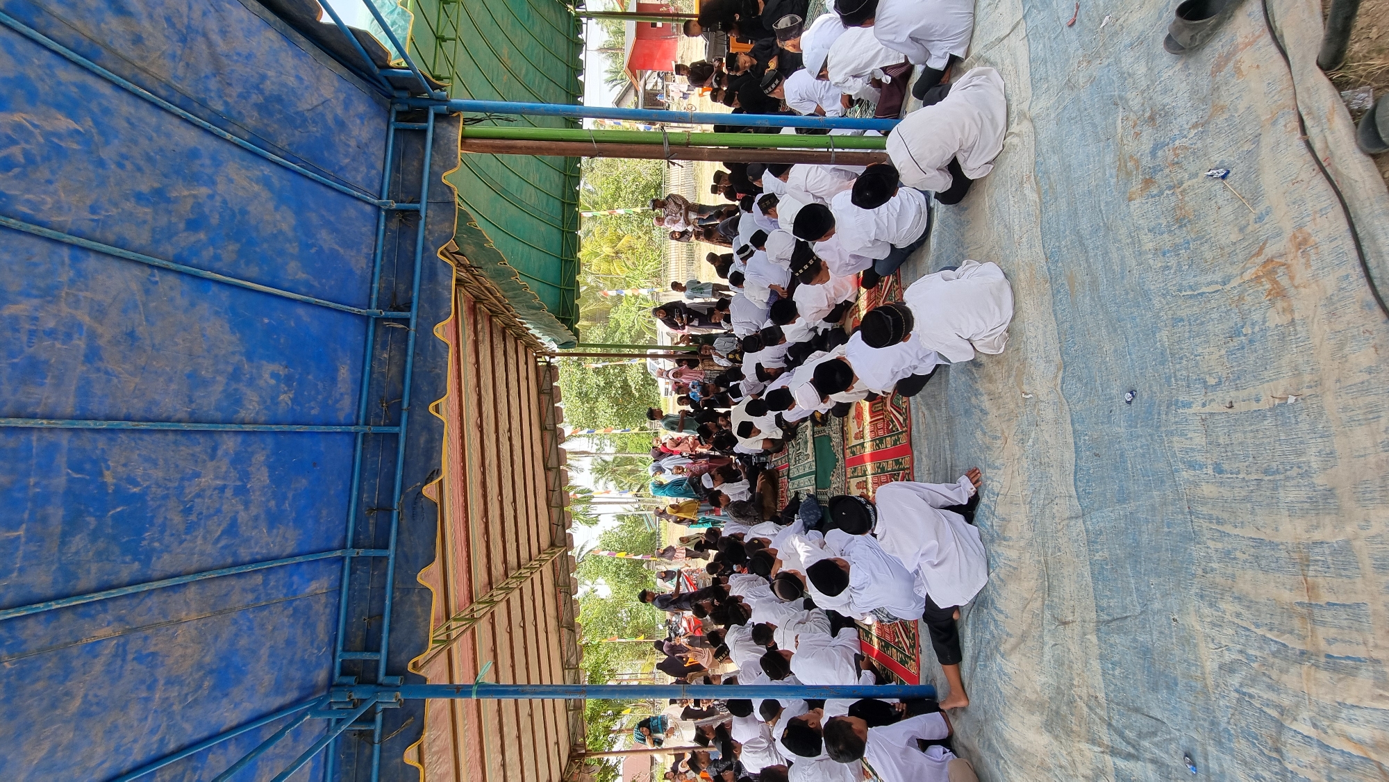 A group of people, predominantly young boys in white attire, are gathered under a blue and green tarp. The setting appears to be outdoors, likely in a community area, as indicated by the presence of trees and a building in the background. The ground is covered with a large blue tarp, and some are sitting on prayer mats. A few adults are visible among the children, some appearing to lead or supervise. The overall atmosphere suggests a religious or educational gathering. The image is captured in Tumpokbeurandang, Indonesia.