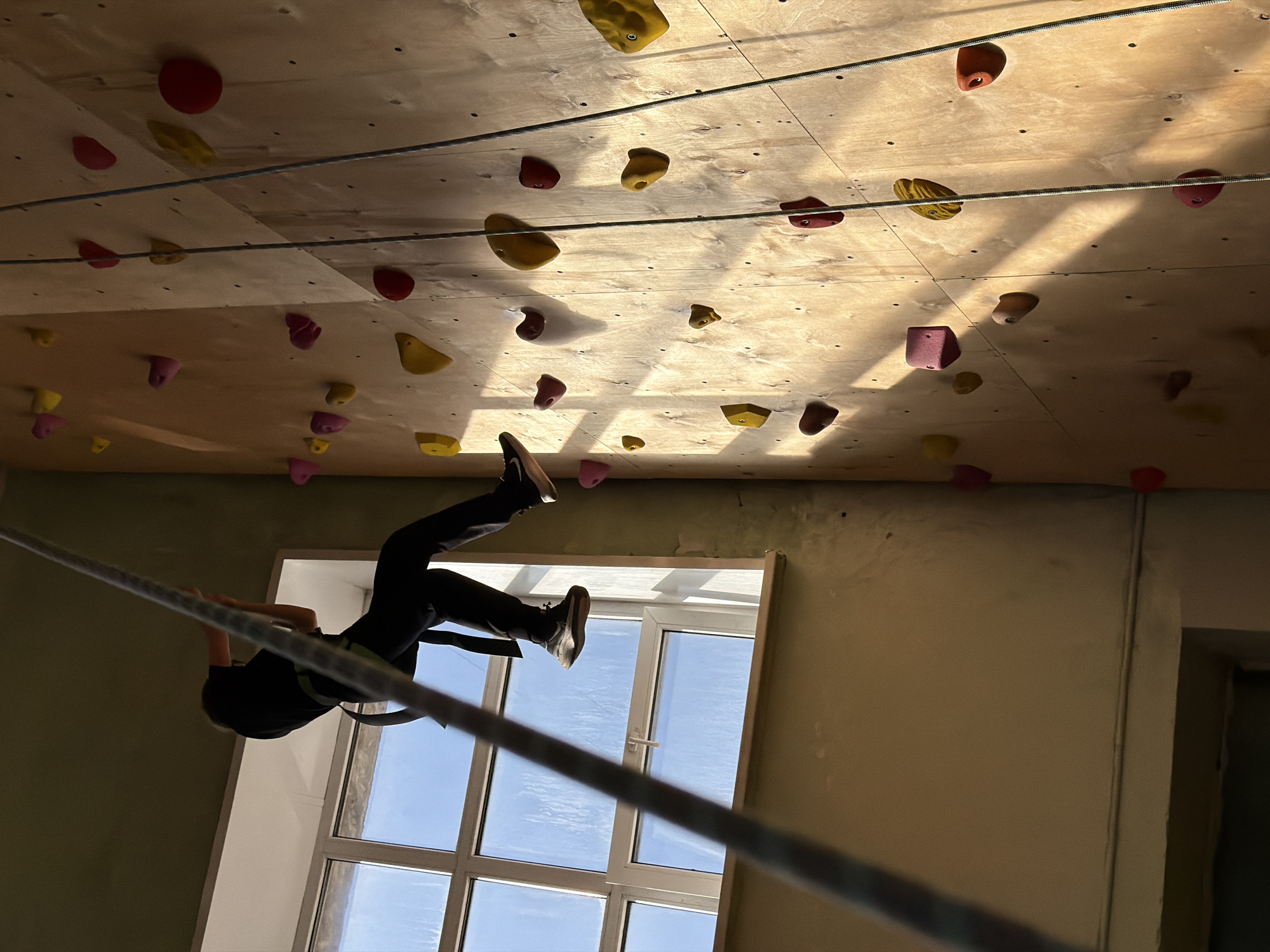 A person is suspended by a rope in front of a climbing wall inside a building. The climbing wall is wooden and covered with various colored climbing holds. A window with a grid pattern is behind the person. The room has painted walls. The location is Izhevsk, Russia.