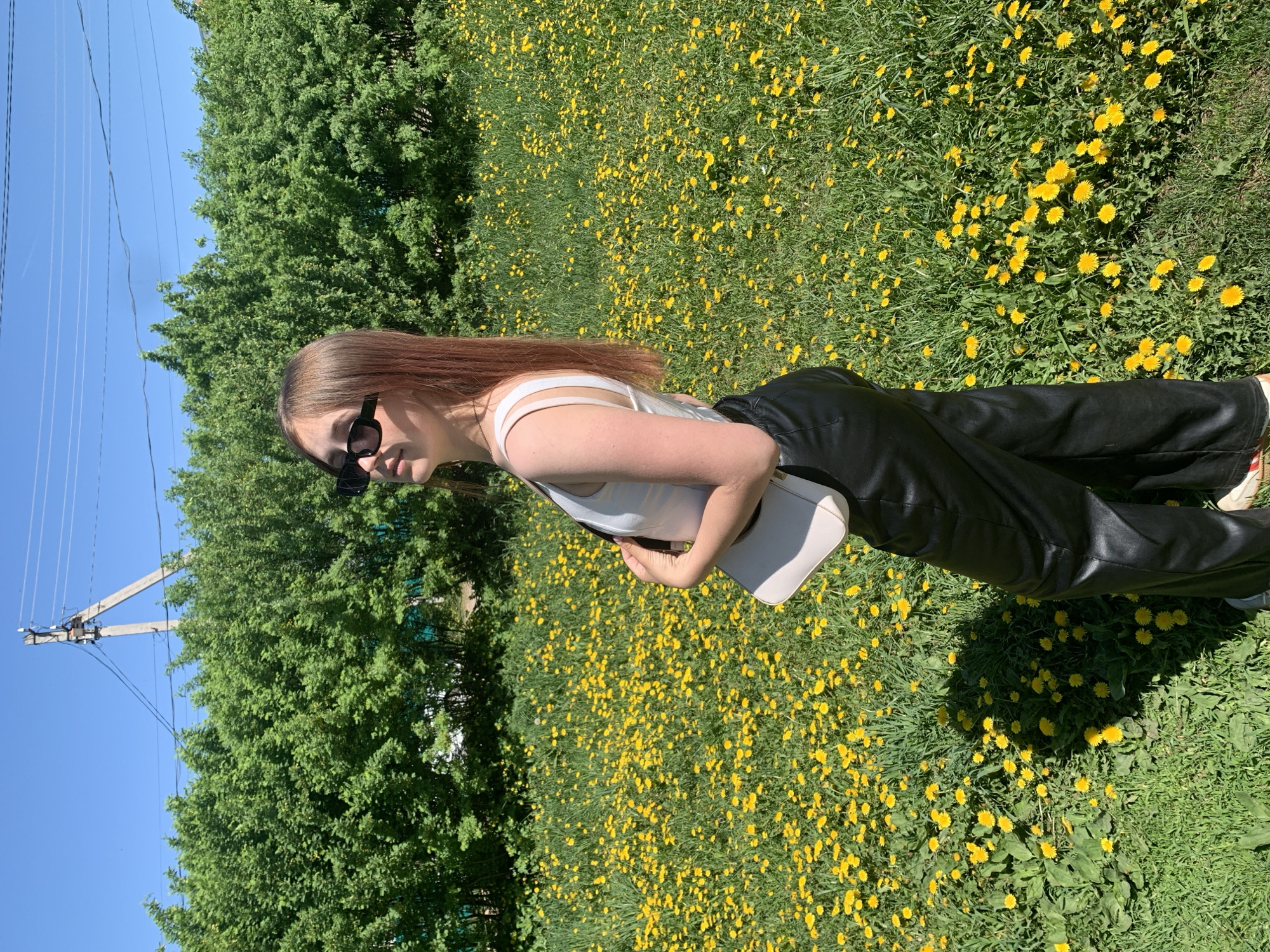 A young woman stands in a field of dandelions on a sunny day in Фоки, Russia. She is wearing a white tank top, black leather pants, and sunglasses. She is holding a white purse. The background includes a green tree and a blue sky with a utility pole.