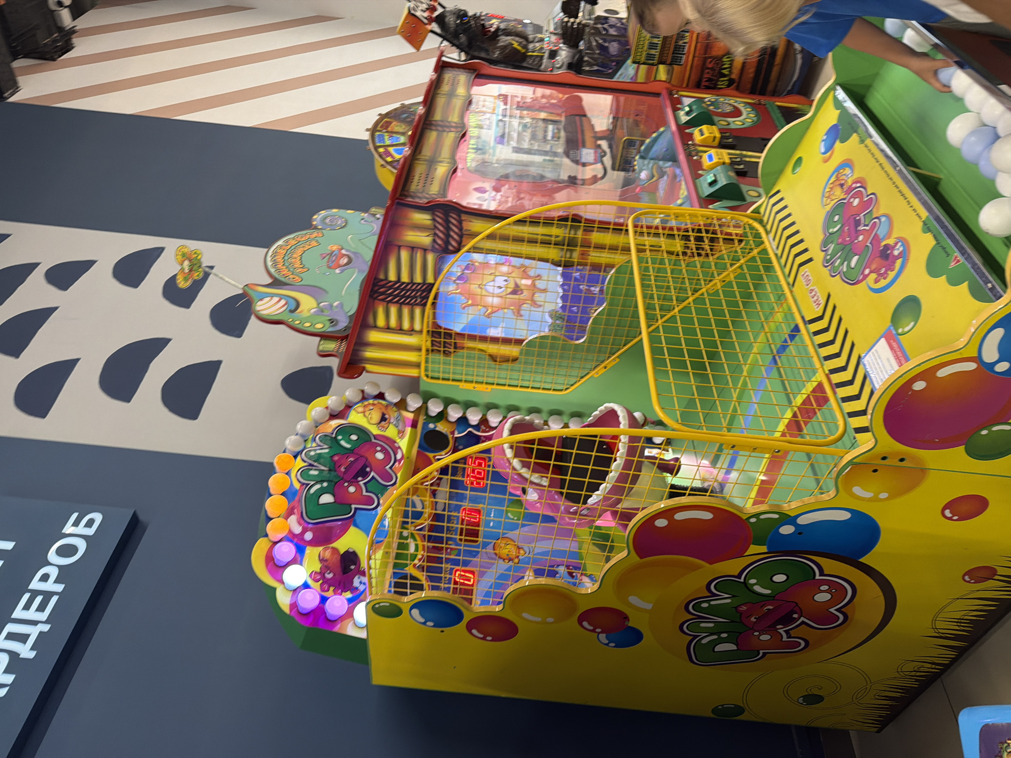 A yellow arcade game with a pink dinosaur mouth and a yellow net is in the foreground. The game has colorful graphics featuring bubbles and the text "DINO POP". Behind it, another arcade game with a tropical theme is visible, featuring a sun character and a gorilla. In the background, a wall is decorated with a pattern of dark blue half-circles on a white background. The scene is indoors, likely in an arcade or entertainment center. A child's arm is visible reaching towards a basket of white and light blue balls at the bottom of the yellow arcade game.