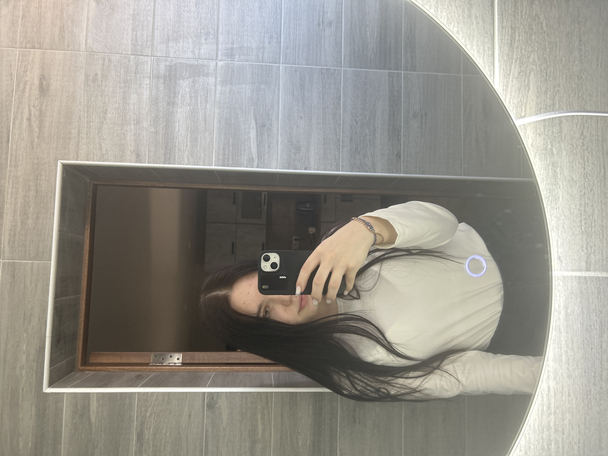 A woman takes a selfie in front of a mirror in a bathroom. The woman has long, dark hair and is wearing a white long-sleeved shirt and dark pants or a skirt. She is holding a black iPhone in her right hand. The bathroom has gray tiled walls and a large, oval mirror with LED lighting around the edge. The mirror reflects a brown wooden cabinet or door. There is no indication of the location being Kemul, Russia.