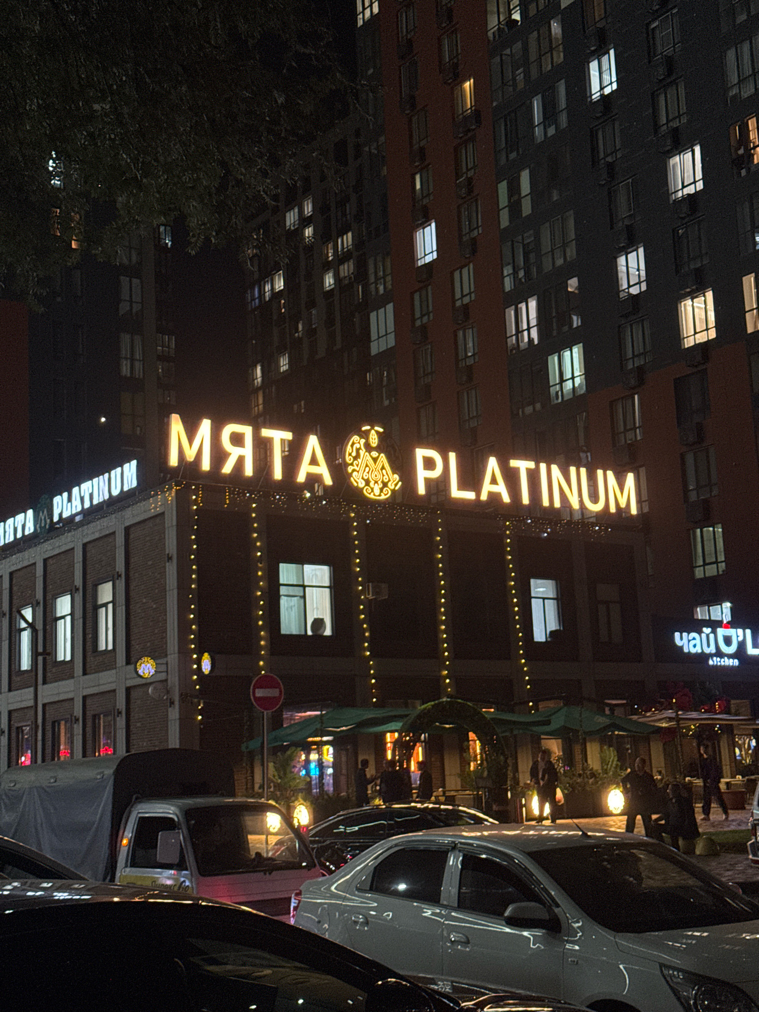 In Almaty, Kazakhstan, at night, a multi-story building with illuminated windows is visible. The facade of a lower building below features prominent illuminated signs. The largest sign reads "МЯТА PLATINUM" in large, yellow, capital letters. To the left, another sign reads "MЯTA PLATINUM" in white, capital letters. Decorative strings of small, yellow lights hang from the edge of the lower building's roof. Several cars are parked or moving in front of the building, including a white truck with a covered bed and a white sedan in the foreground. People are visible on a street or sidewalk area in front of the building, near the entrance to a restaurant or establishment with green awnings. Another illuminated sign, reading "чай O'L" with "Kitchen" below it, is also visible on the right side of the frame. A "no entry" sign is mounted on a pole in front of the building. The overall scene depicts a lively urban environment at night with businesses illuminated and vehicles and pedestrians present.