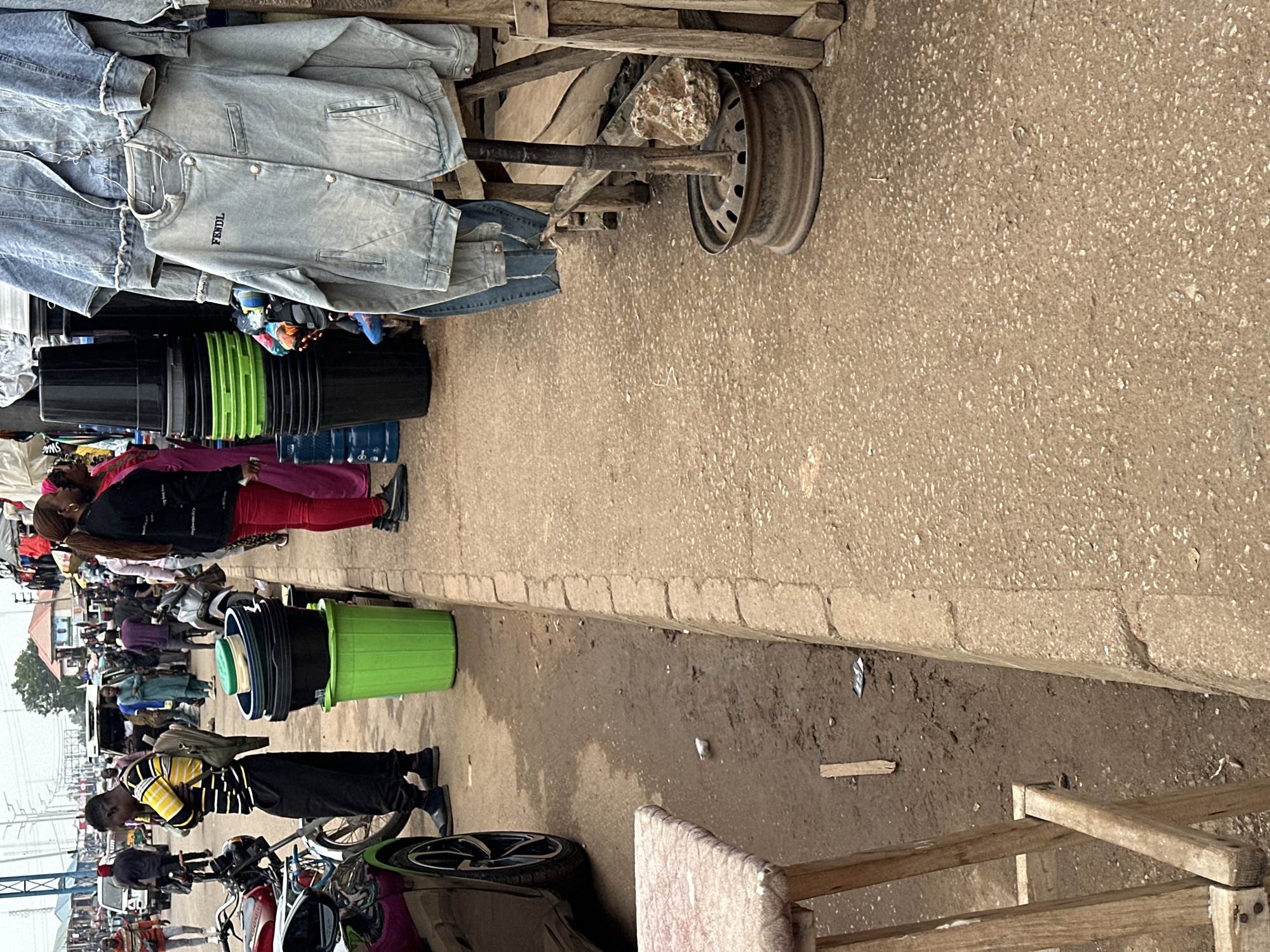 The image depicts an outdoor scene at a market in Bukuru, Nigeria. A man wearing a yellow and black striped shirt and black pants stands with his arms crossed, looking down at the ground. He has a backpack slung over his left shoulder. To his right, a stack of large black plastic buckets sits on top of a bright green plastic bucket.
Behind the man and the buckets, several people are walking in the background. A woman in a black jacket and red pants stands to the right of the buckets, facing away from the camera. She is standing next to another woman wearing a pink garment.
Further back, various stalls are visible, with clothing, including denim jackets, hanging from displays. A motorcycle is parked to the left of the man. The ground appears to be a mixture of dirt and pavement, with some debris scattered around. A wooden stool with a patterned seat is visible in the lower left corner of the image. The overall impression is of a bustling marketplace.