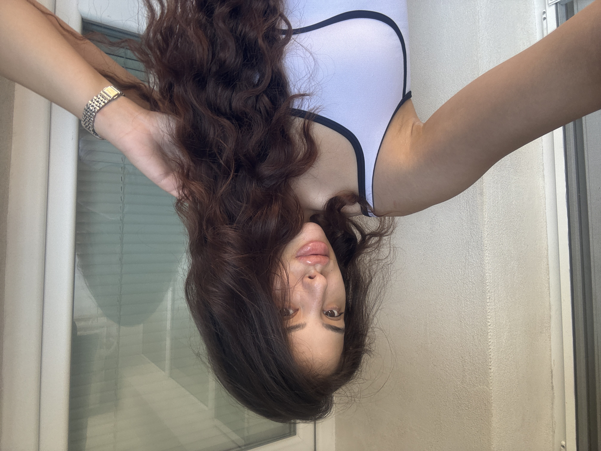 A woman with long, dark, wavy hair and fair skin is pictured from the chest up, holding her hair with her right hand. She is wearing a white tank top with black trim. She has on a watch on her left wrist. The background is a textured, light-colored wall to the left and a window with blinds to the right. The overall lighting suggests an outdoor setting during the day, possibly a balcony or patio. There is no indication of the location being the city of Plovdiv, Bulgaria.