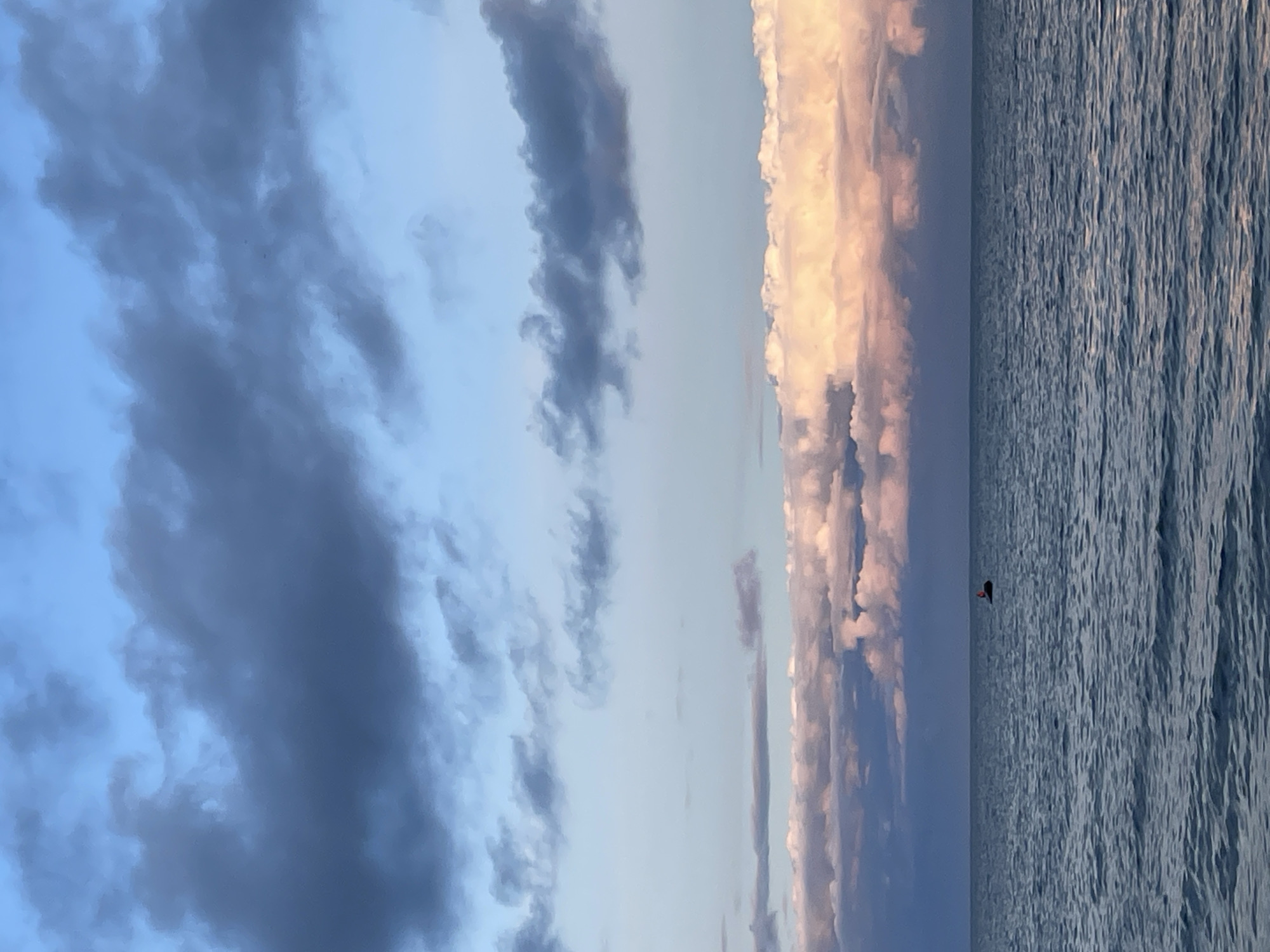 The image depicts a tranquil seascape, likely at sunset or sunrise, given the soft hues in the clouds. The sky is a gradient of pale blue and soft pink, with layers of darker, textured clouds in the upper portion of the frame. Below the sky, a band of fluffy, illuminated clouds is visible, tinged with the warm colors of the setting or rising sun. The ocean occupies the lower half of the image, its surface rippled by a gentle swell. A small, dark object, possibly a buoy or a small boat, is visible in the middle distance on the water. The horizon line is clearly defined, separating the sky and sea. The scene evokes a sense of calm and serenity. Based on the provided context, this seascape is located in the city of Zelenaya Roshcha, Russia.