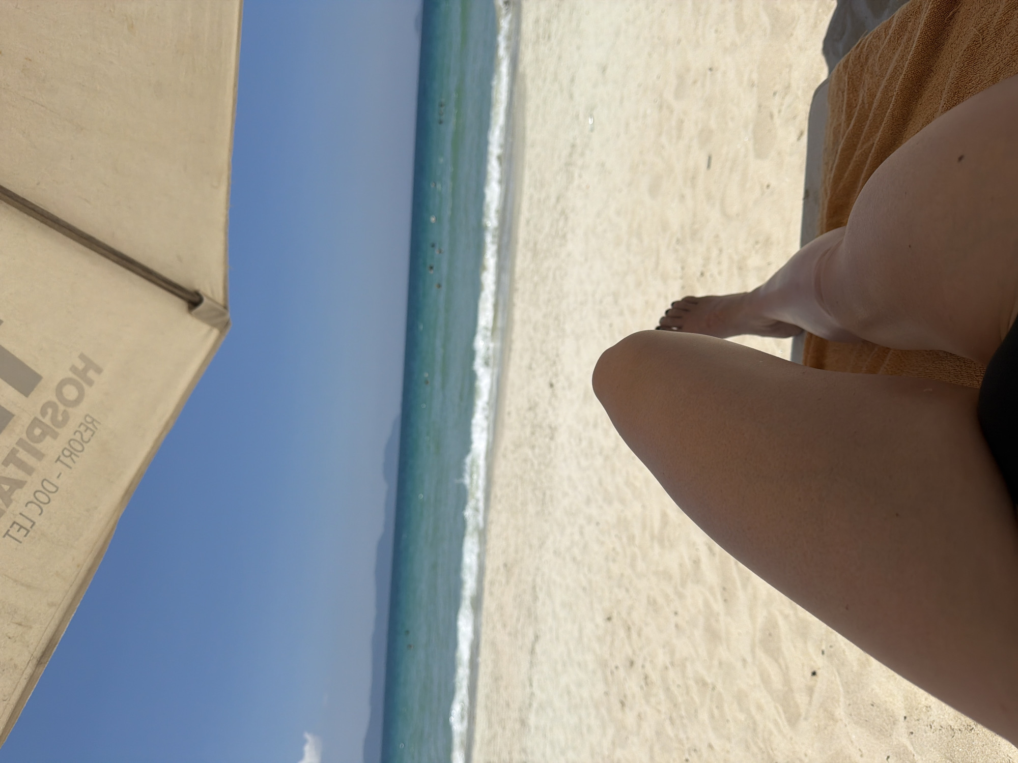 The image presents a first-person perspective of a sunny beach scene in Dong Ninh Hoa Ward, Vietnam. The upper portion of the frame is occupied by the underside of a beige beach umbrella, displaying mirrored text that reads "HOTEL" and "RESORT - DOC GEL". Below the umbrella, a clear blue sky extends to a distant horizon, where a line of low-lying landmasses is visible. The middle ground features turquoise ocean water with small white waves breaking on a light-colored sandy beach. Several dark, indistinct figures are scattered across the water, indicating people swimming or wading. In the immediate foreground, the bare, tan-skinned legs and feet of an individual are prominent. The left leg is bent, while the right leg is extended and rests on an orange-brown towel draped over a beach lounger, which is partially visible at the bottom right. The feet display dark painted toenails. The sand in the foreground is light-colored and shows minor undulations.