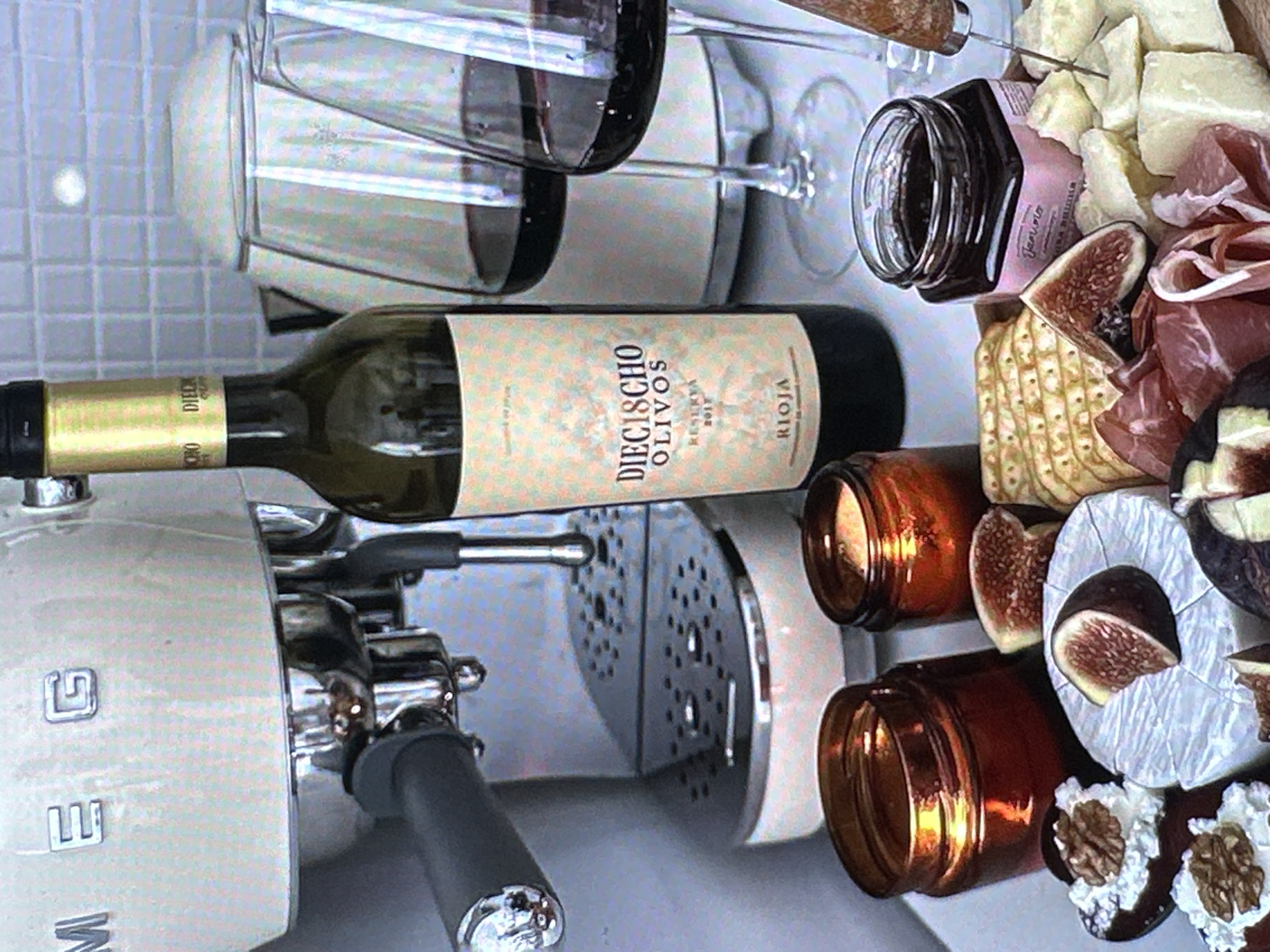 The image depicts a charcuterie board featuring an assortment of food items including cheese, figs, crackers, prosciutto, jam, and chocolate treats with walnuts. A bottle of "DIEGISCHO OLIVOS" Rioja wine and wine glasses are also present. A Smeg coffee maker is visible in the lower left corner. The setting is likely indoors. There is no information available in the image to suggest a location in Yerevan, Armenia.