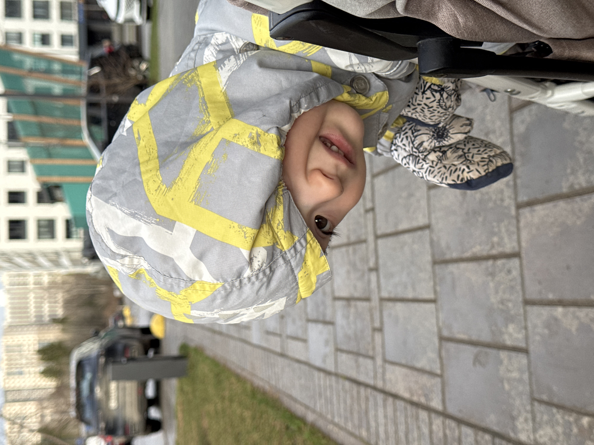 The image, taken in Izhevsk, Russia, captures a happy baby smiling and looking upside down from a stroller in an outdoor, urban setting during daytime. The baby is wearing a grey and yellow patterned hooded jacket and patterned cream and black mittens or booties. The stroller's frame is visible in the upper right. The background shows a paved sidewalk, a strip of green grass, and blurred urban elements including buildings and a vehicle. The lighting suggests an overcast day. No text is visible.