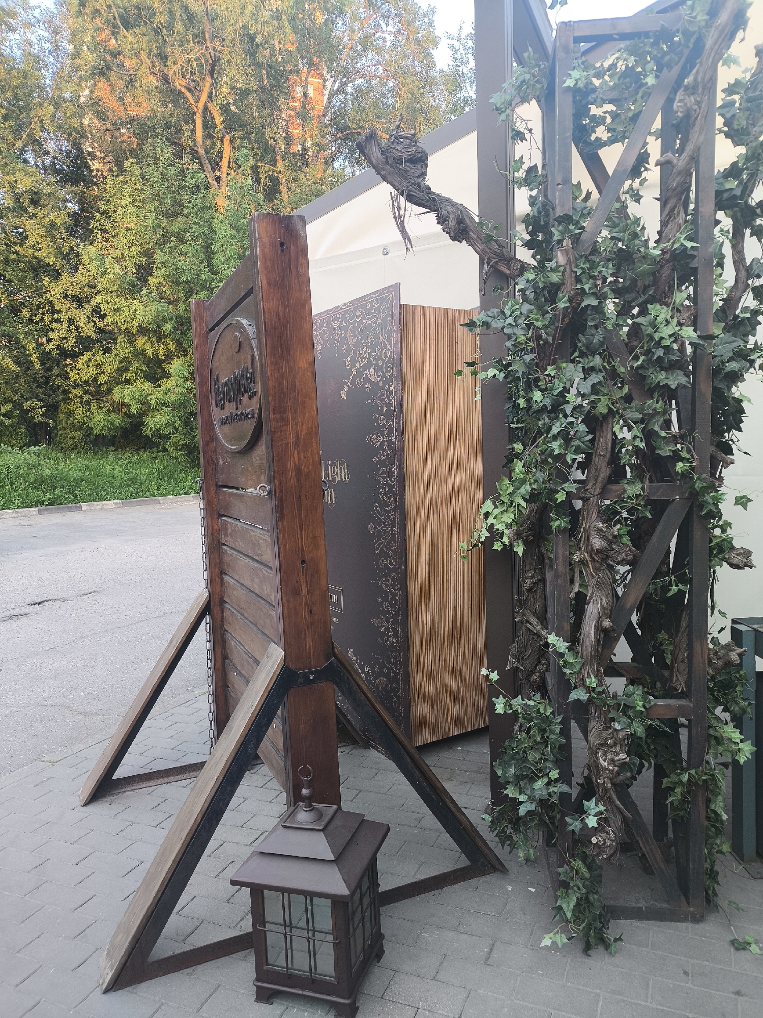 A decorative outdoor display setup is captured in warm, late afternoon or early evening light. In the foreground, a large, dark brown wooden structure stands on a paved surface. It consists of a two-sided stand; one side features vertical wooden planks and a circular plaque, while the other is a solid dark panel with intricate patterns. This stand is supported by sturdy wooden braces that angle outwards onto the pavement. A dark metal lantern with glass panes rests on the ground near the base of these supports.
To the right of the wooden stand, a tall, dark wooden trellis or frame is heavily adorned with artificial ivy and gnarled, intertwining branches, giving it a rustic or fantastical appearance. Behind these structures, a white building or large tent is partially visible. The background is dominated by lush green trees and foliage, illuminated by the golden sunlight, with a clear sky peeking through. Beyond the paved area, an asphalt road or pathway is visible.
Visible text on the circular plaque of the wooden stand appears to be in Cyrillic script, possibly "Кукушка!" or similar, with another word, possibly "РЕСТОРАН" (Restaurant), below it. On the larger dark panel, partially obscured English text reads "light" and "in", and "TH" is visible near the bottom edge. No people are present, and the scene suggests an entrance or decorative element for an establishment or event.