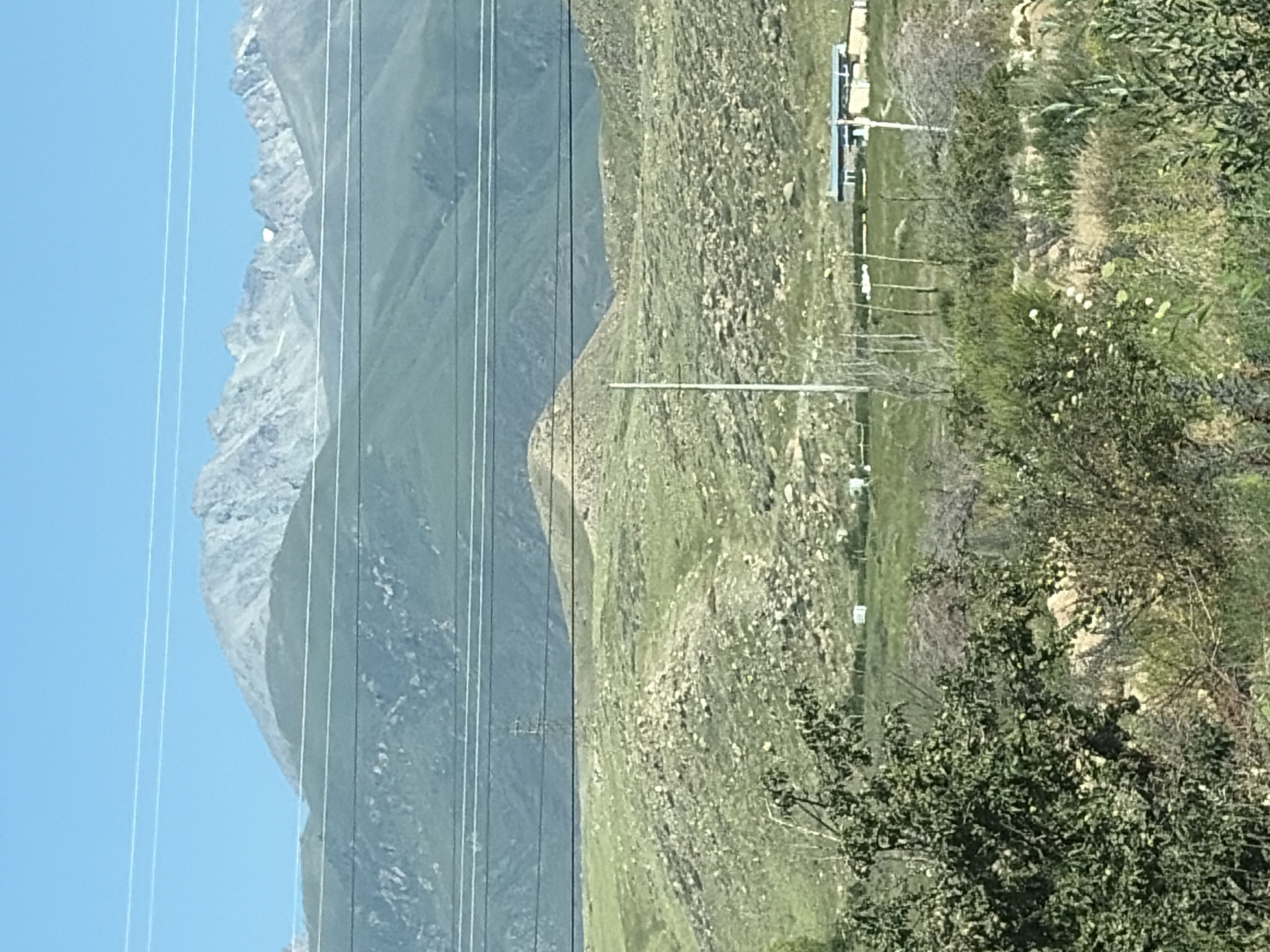 The image depicts a mountainous landscape under a clear blue sky. In the foreground, there are several trees and bushes with green foliage, and some dry brush. A few white structures, possibly tents or small buildings, are visible in the middle ground, along with a long, low building with a blue roof. A tall, slender utility pole stands near the center of the frame. In the background, rolling green hills are covered in rocks and vegetation, leading up to imposing, rocky mountain peaks. Several power lines are strung across the scene, obscuring parts of the landscape. The overall scene appears to be a rural or natural environment, possibly in a mountainous region. There are no people visible in the image.